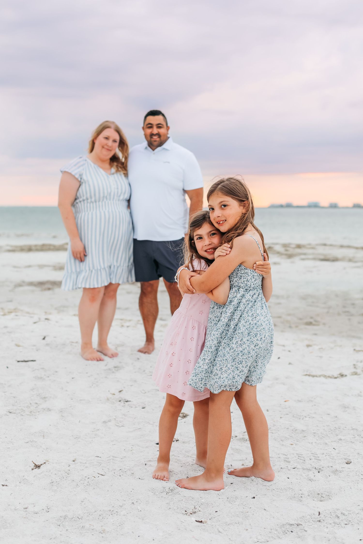 Family of four poses on a sandy beach at sunset, two girls hugging in front while parents stand smiling behind them.