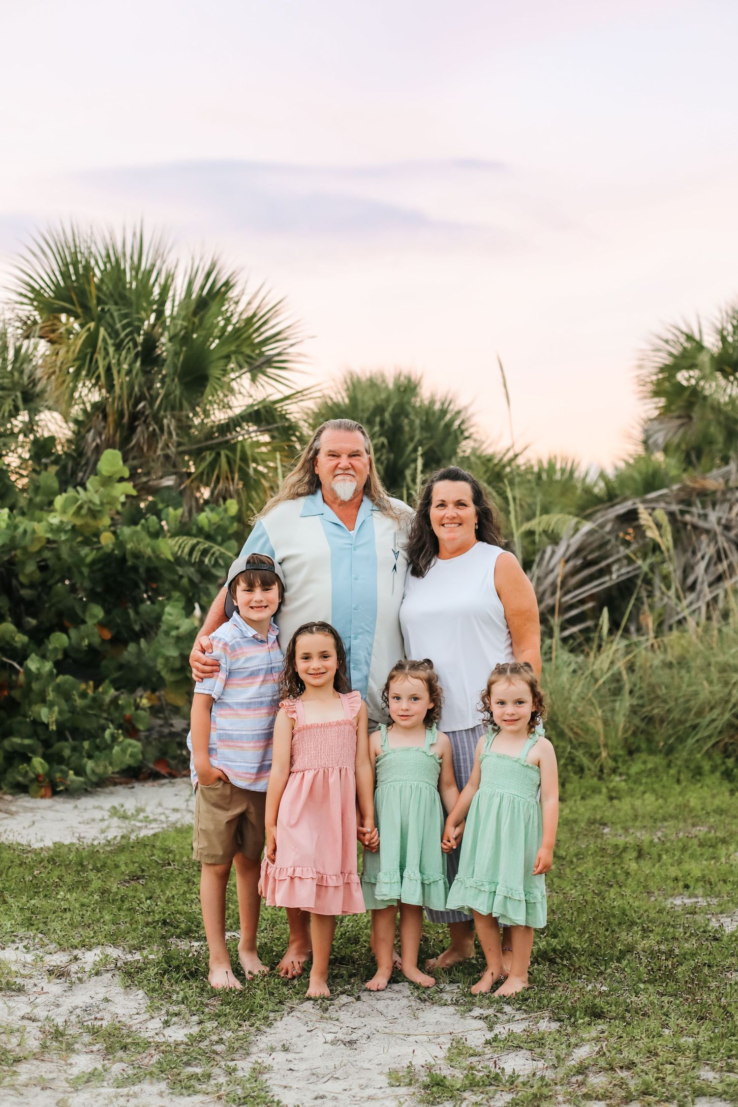 Family of six poses barefoot on sandy beach path surrounded by palm trees at sunset in Florida.
