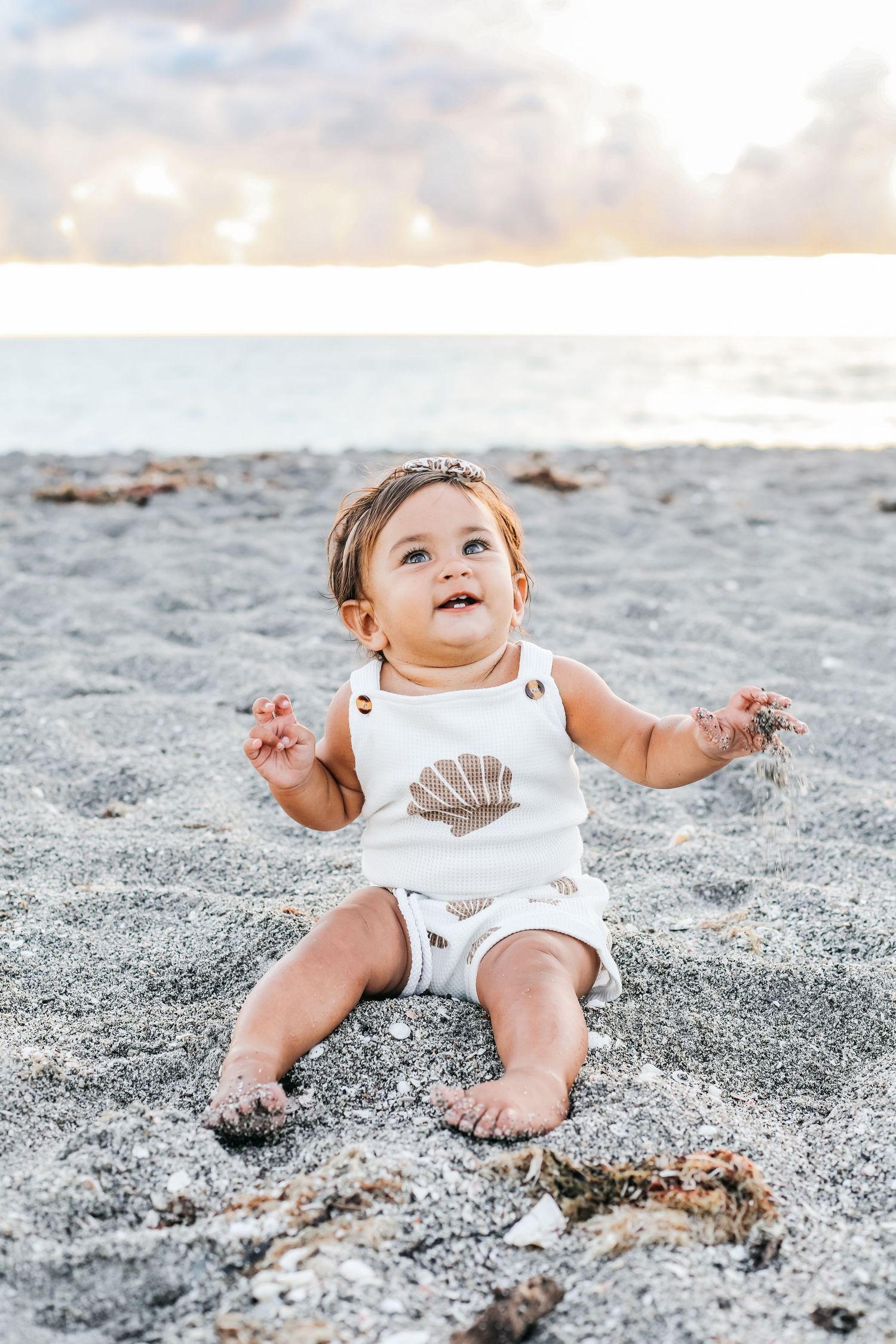 A baby in a white outfit plays and smiles while sitting on sandy beach with seashells scattered around.