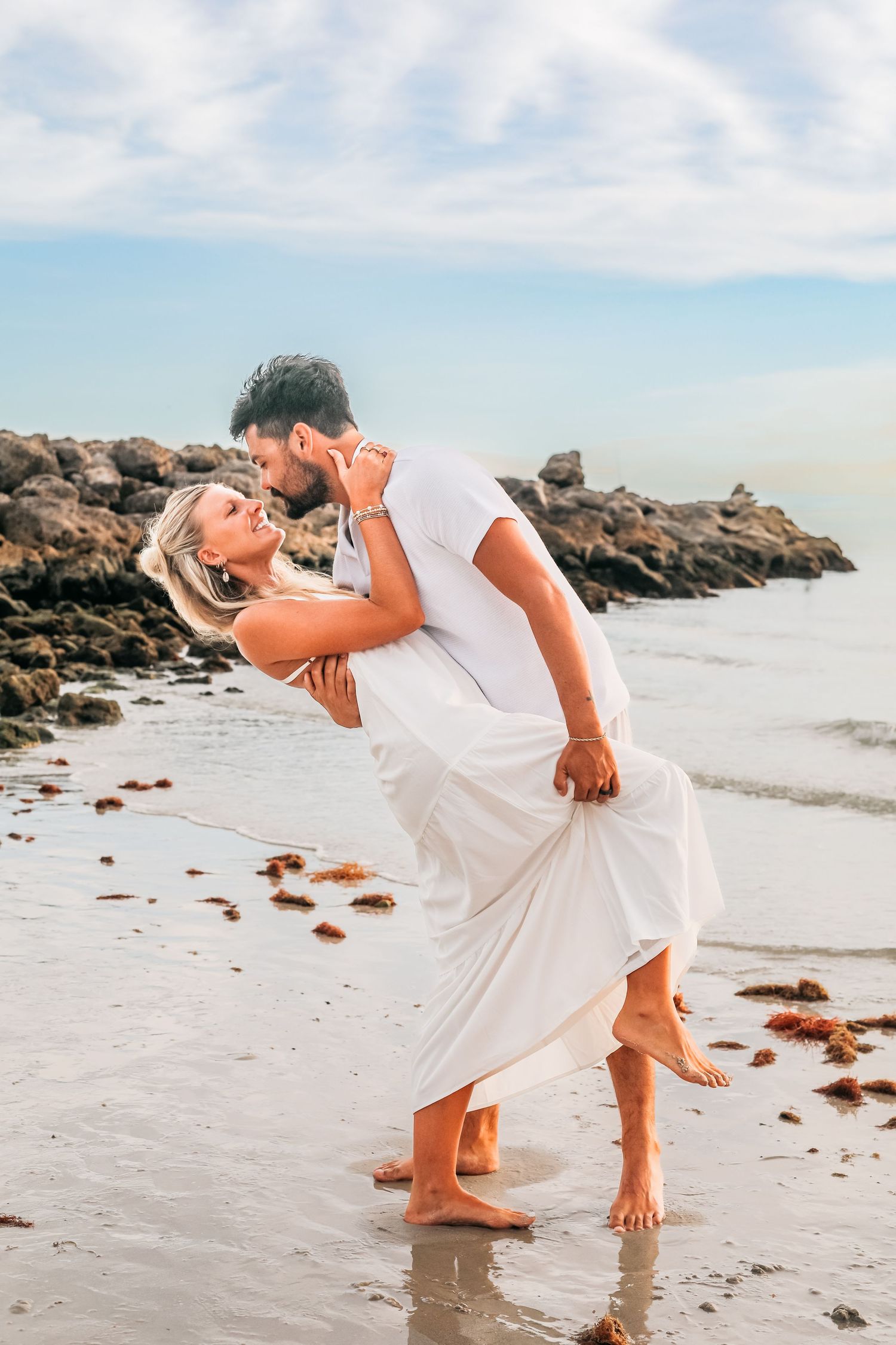 Romantic couple in white clothing dancing on beach near rocky coastline.