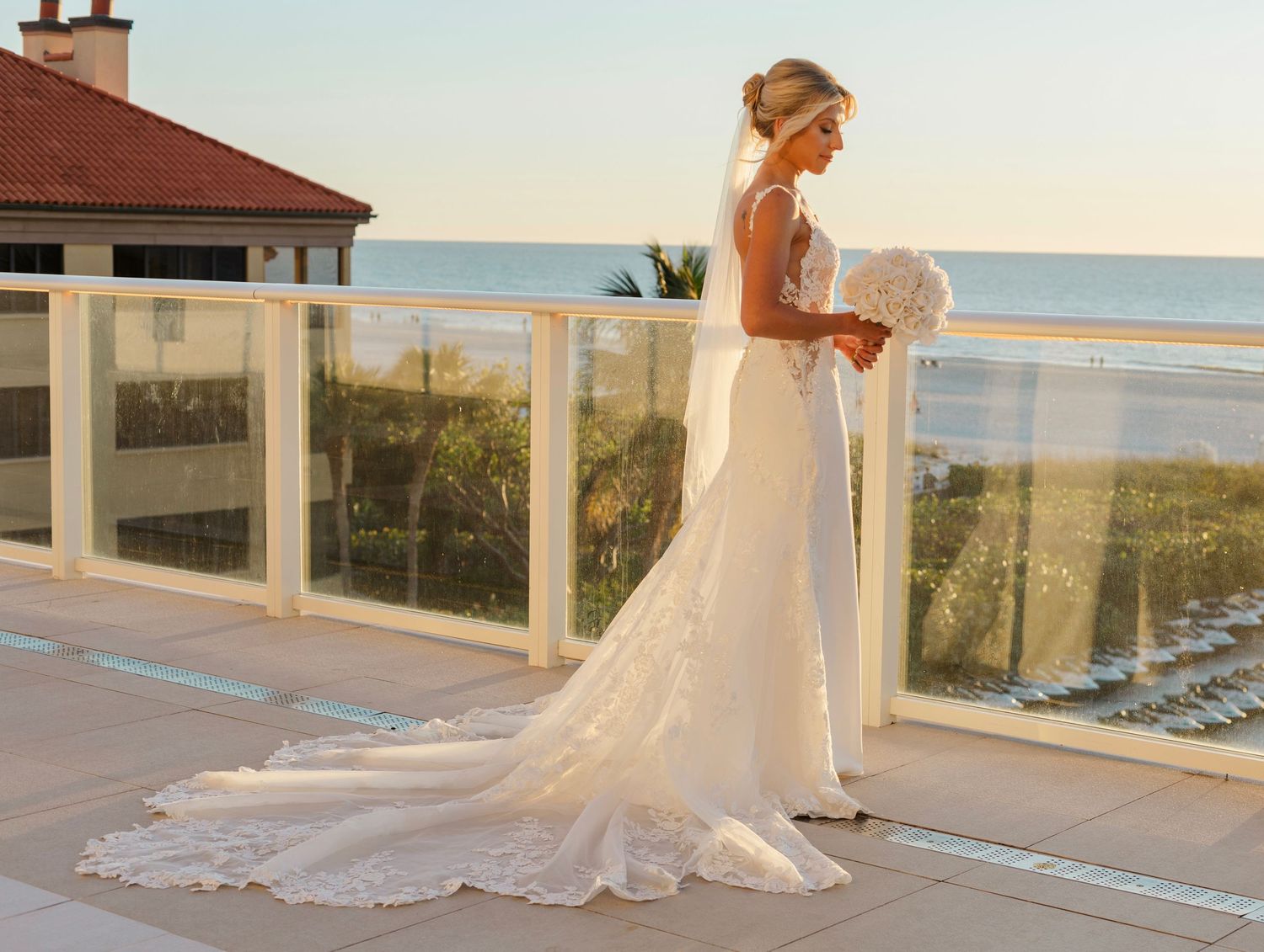 Bride in elegant lace wedding gown with train on oceanview terrace at sunset.