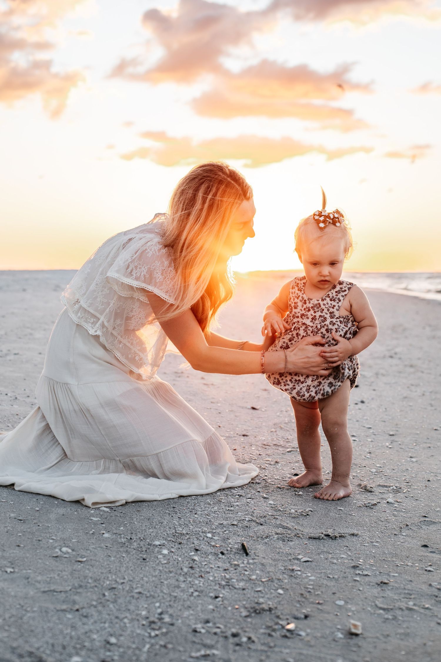 A tender sunset beach moment between parent and child wearing white and floral outfits on the sand.