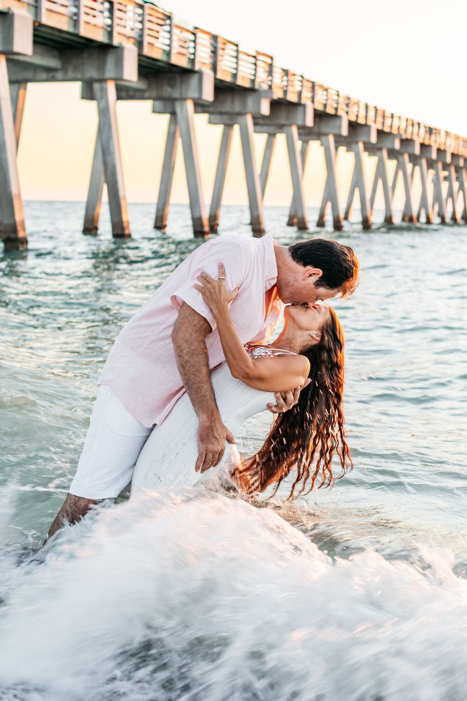 A couple shares intimate moments near a pier as ocean waves crash around them during a beach photoshoot.