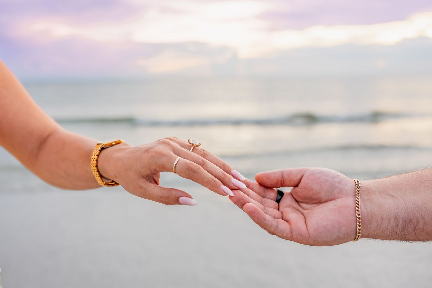 Two hands reaching toward each other on a beach at sunset, adorned with gold bracelets and rings.