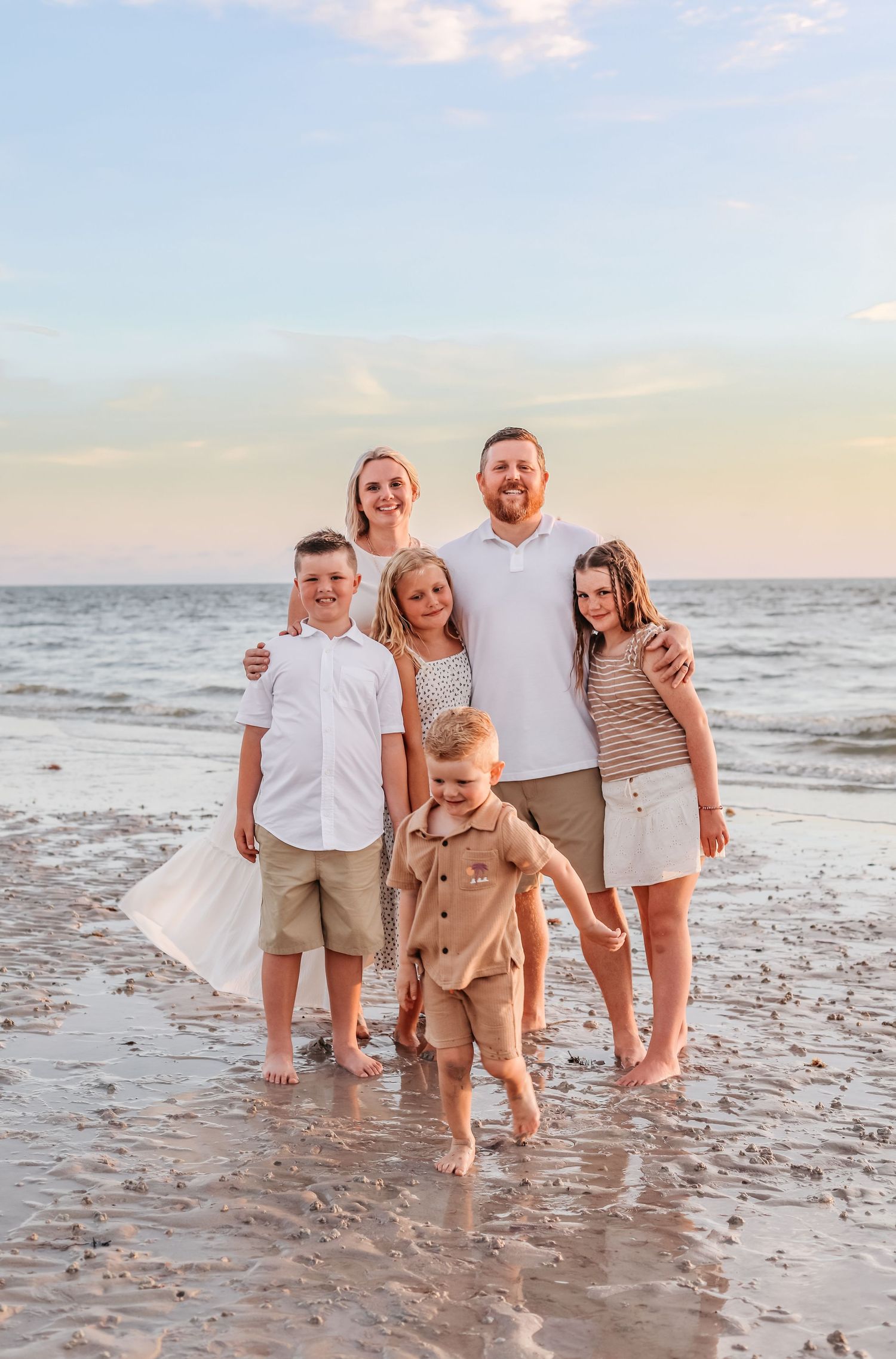 Family of six posing together on a sandy beach at sunset, wearing coordinated neutral and white outfits.