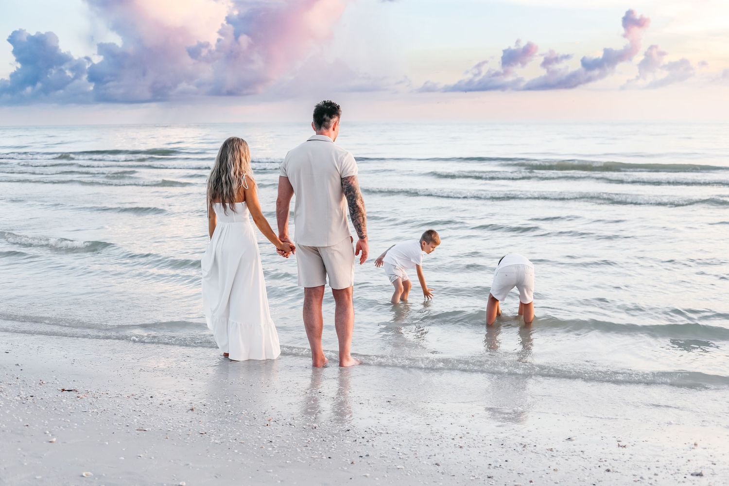 A family wearing white outfits walks along the shoreline during sunset at a beach.