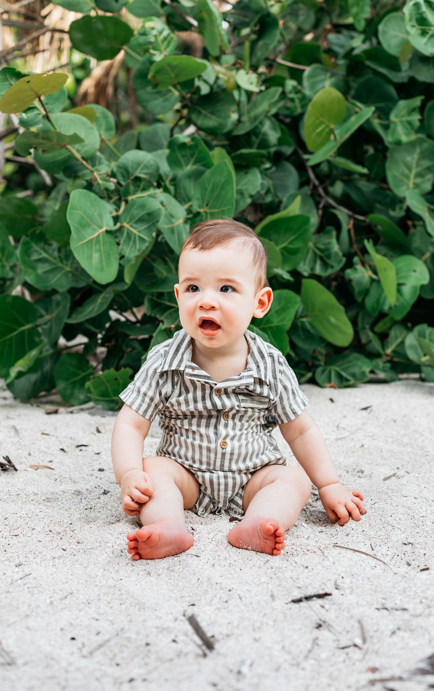 Small baby in striped outfit sitting on white sand with lush greenery backdrop.