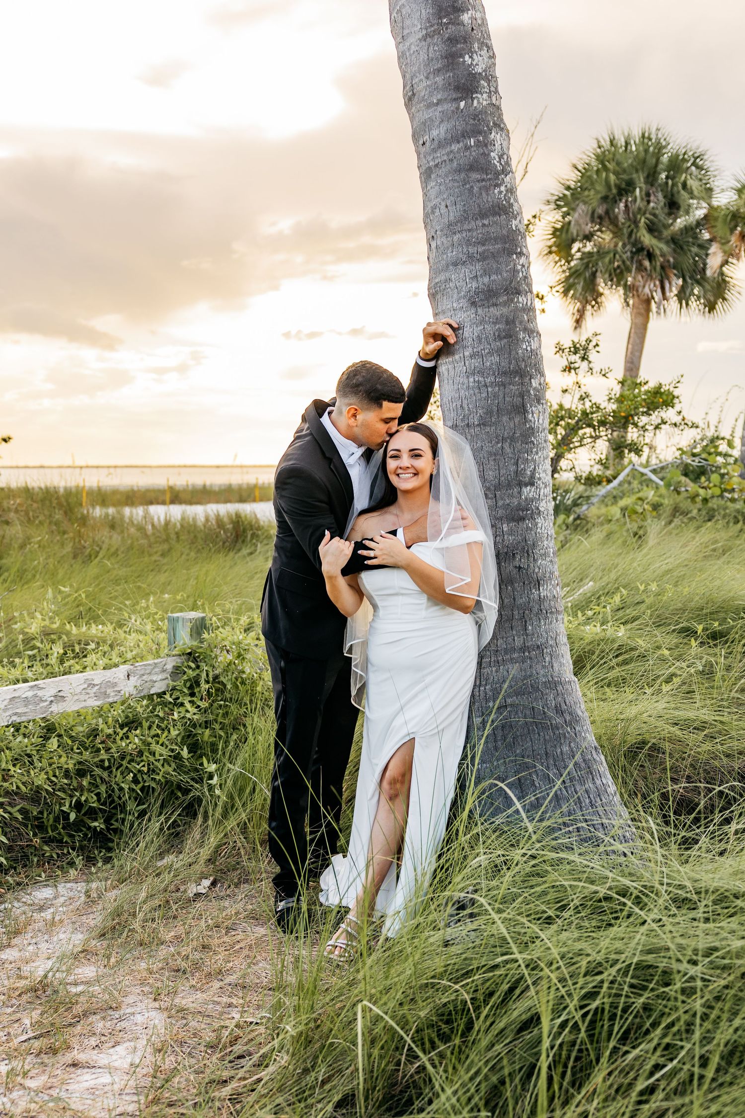 A bride and groom embrace by a palm tree at a beach wedding, smiling joyfully at sunset.