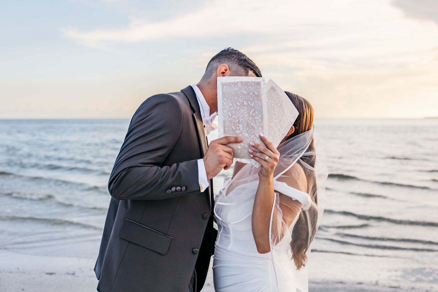 Newlywed couple kissing behind wedding vows paper on a beach at sunset, bride in white gown with veil.