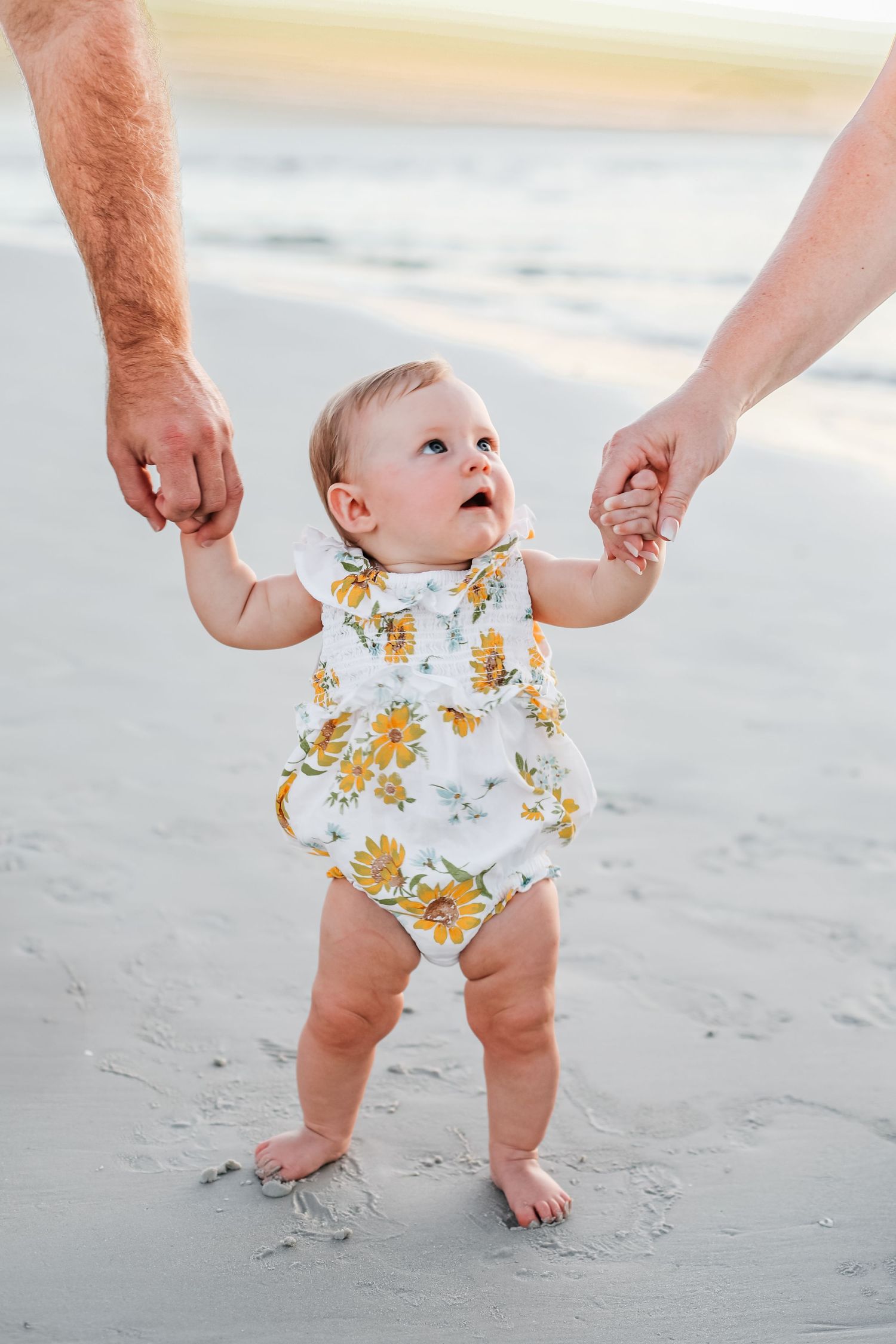 Baby in yellow floral outfit takes first steps on beach holding hands with parents.