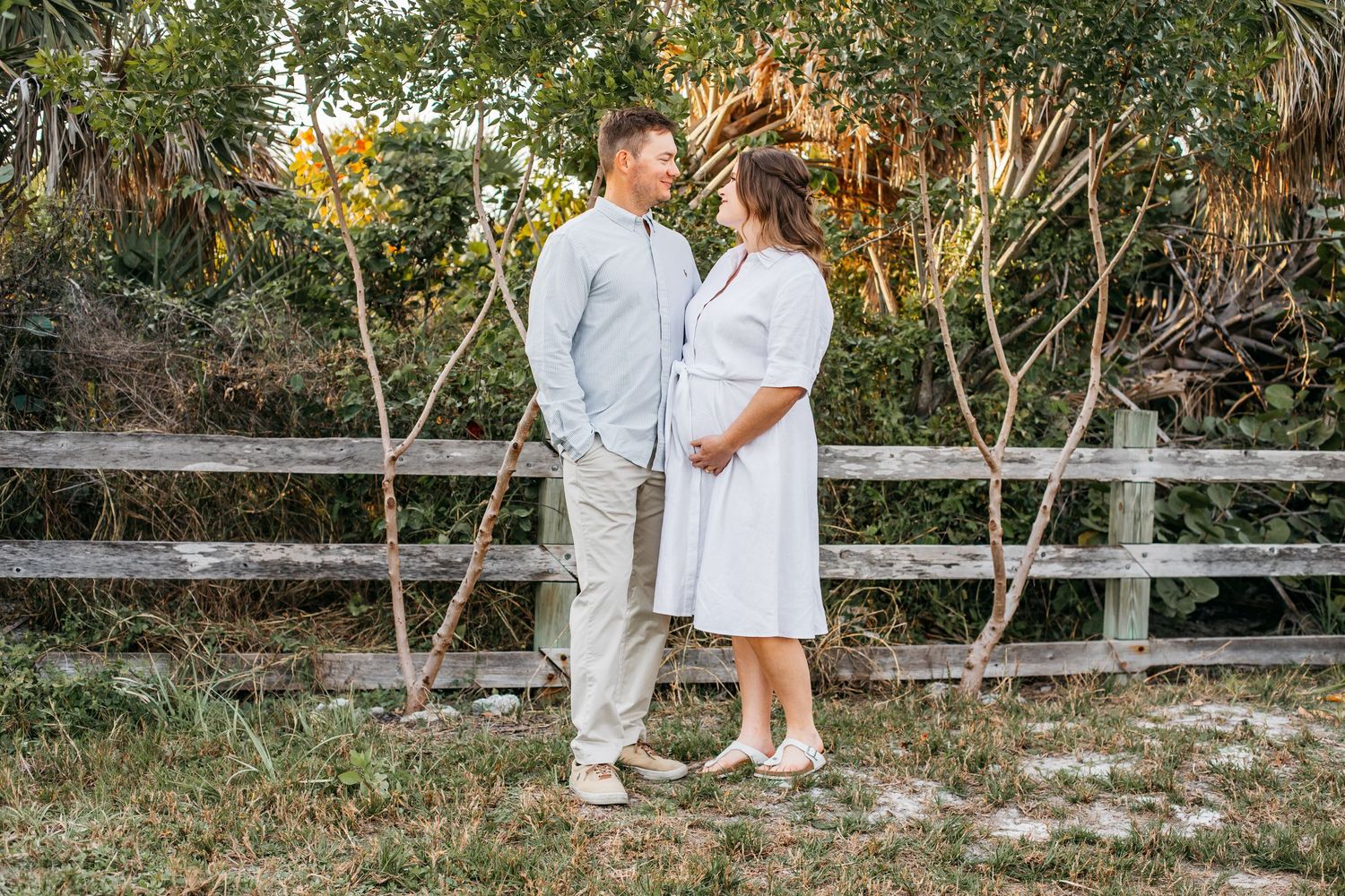 Couple standing together by rustic wooden fence in outdoor setting, woman in white dress, man in light blue shirt.