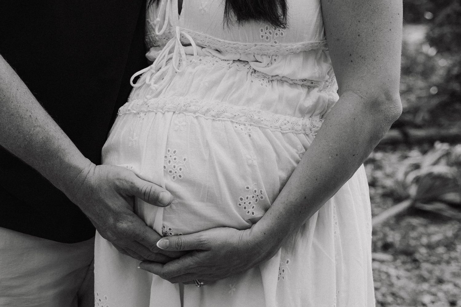 Black and white photo of a pregnant woman in a white dress with her partner's hands cradling her baby bump outdoors.