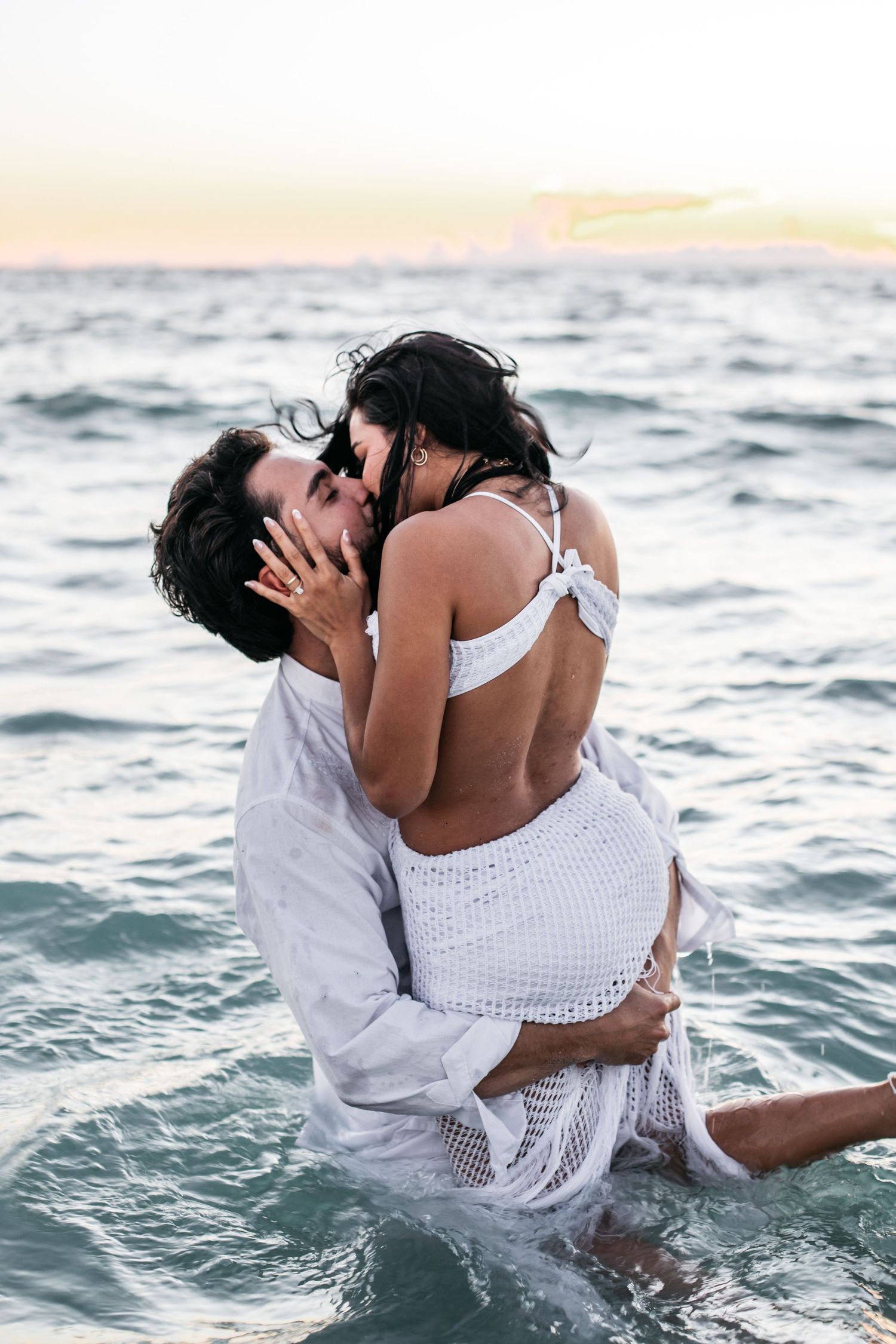 A romantic couple embraces and kisses in the ocean at sunset wearing white outfits.