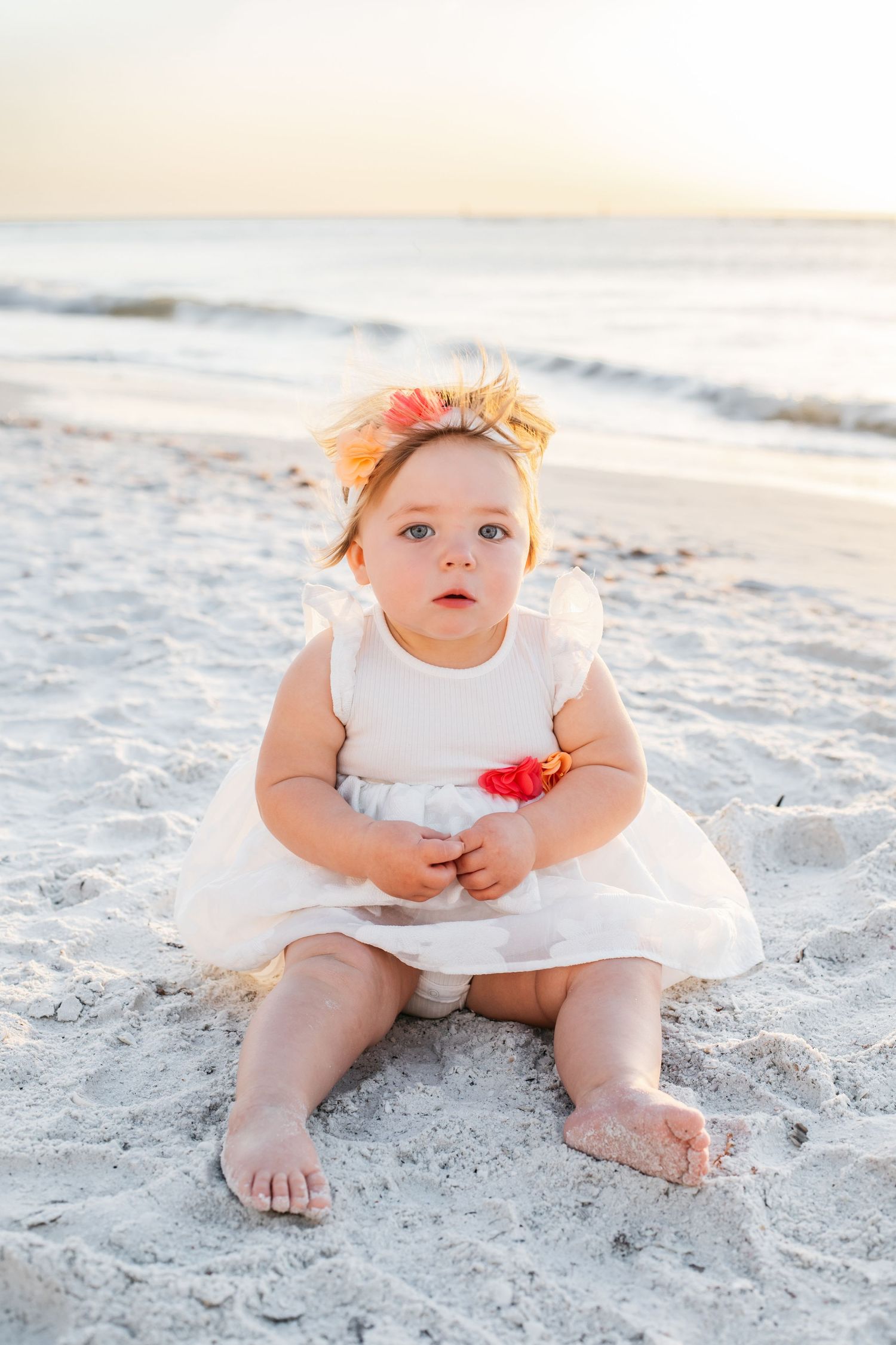 A baby in a white dress sits on sandy beach during golden hour sunset, creating a serene coastal portrait.