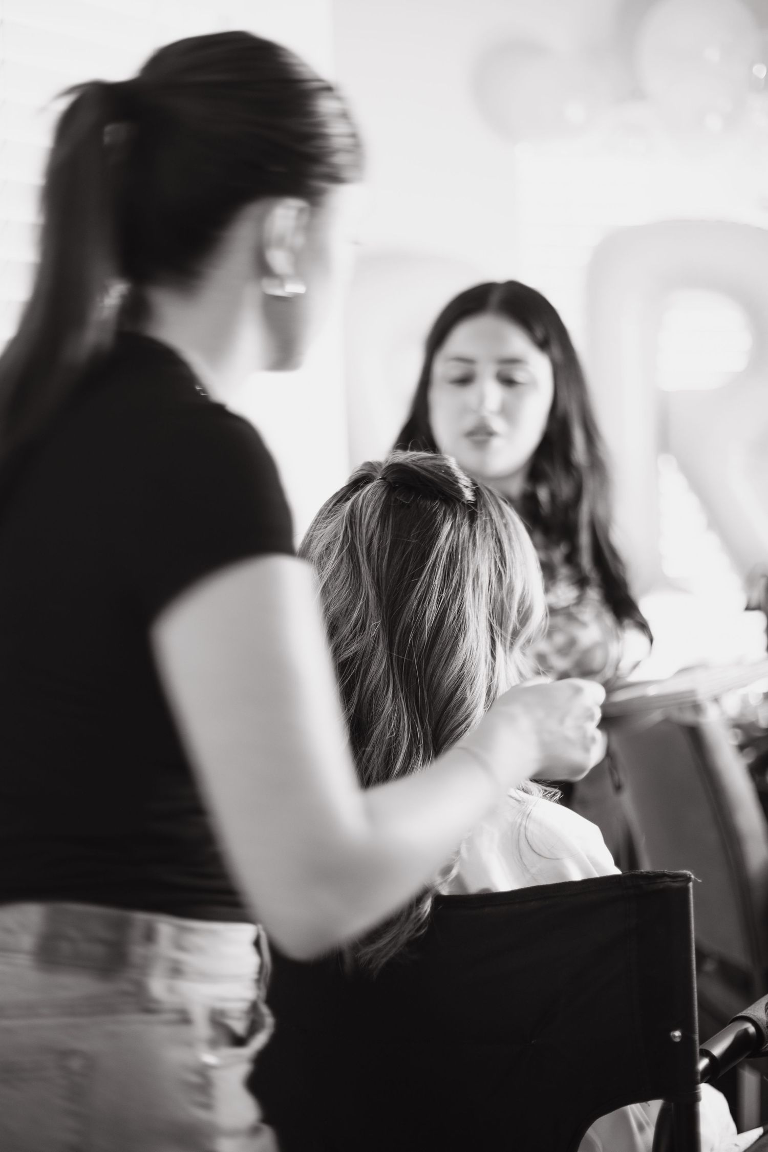 Black and white photo of a hairstylist working on a seated client while another woman watches in a bright room.