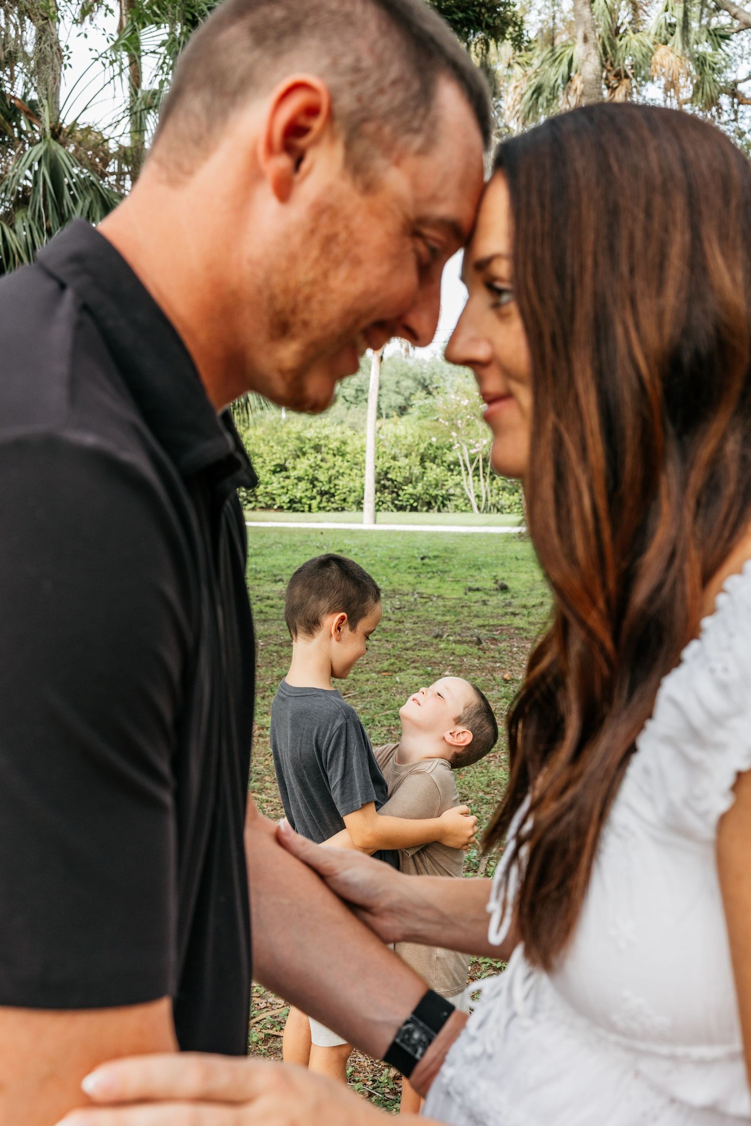 A couple touches foreheads tenderly while their two young sons play together in a lush green park background.