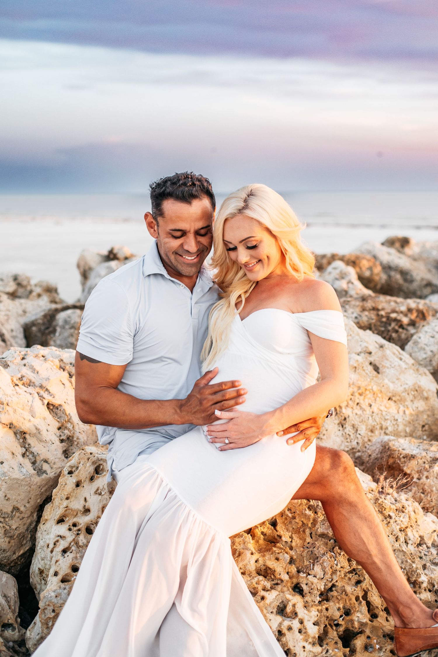 A romantic maternity photoshoot on rocky coastal terrain at sunset with soft pink and purple sky.