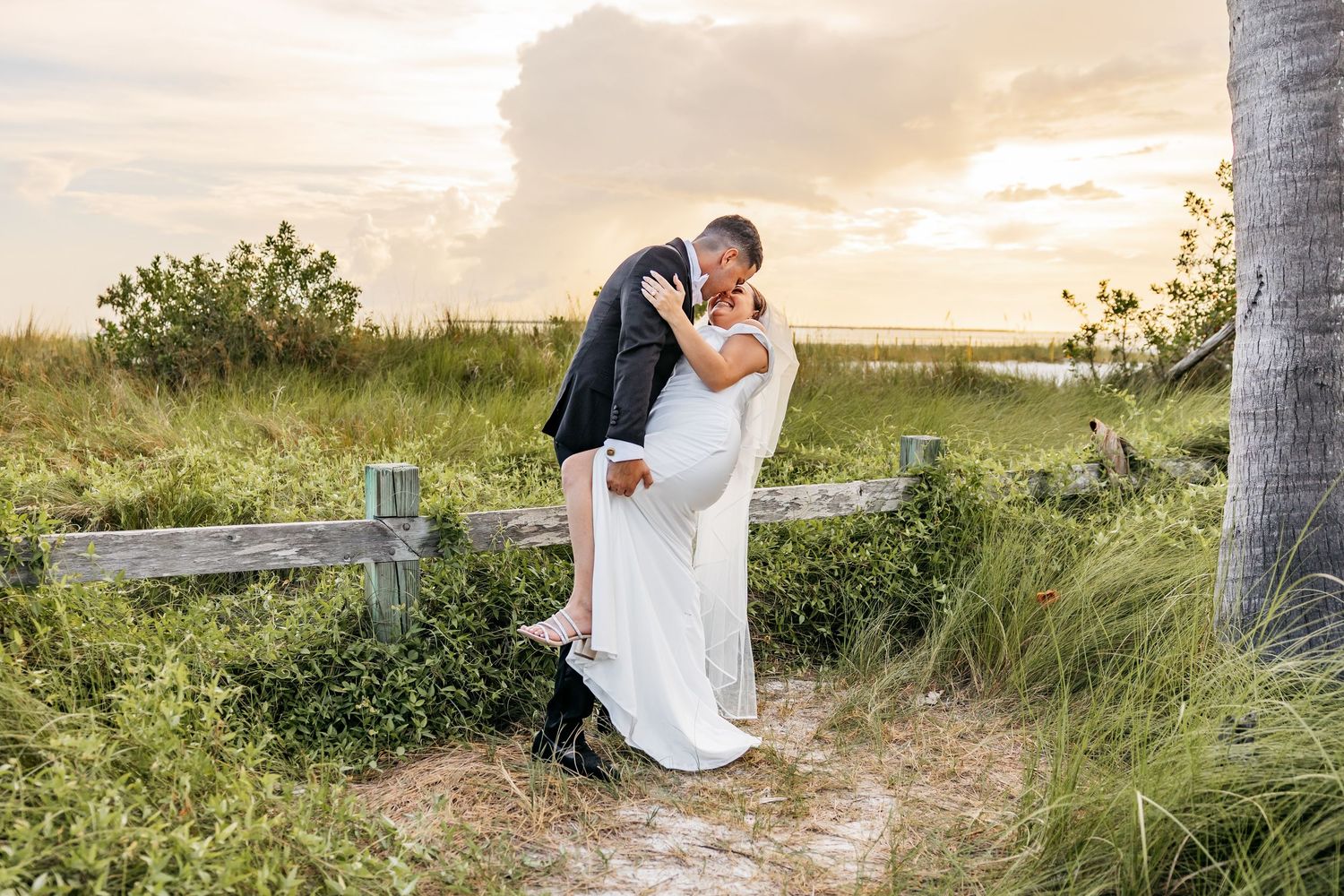 Groom dips bride in romantic sunset kiss on sandy beach path surrounded by lush green grass and palm trees.
