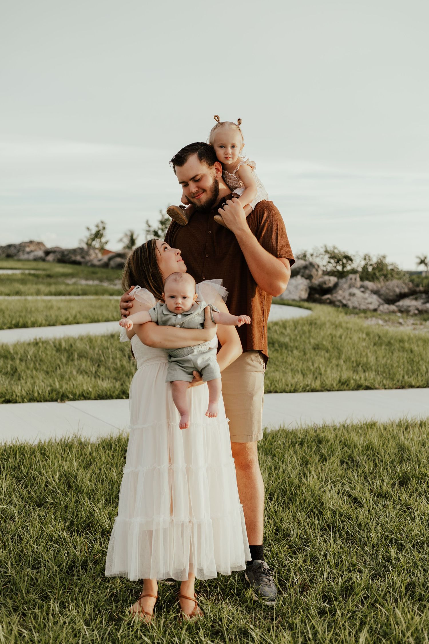 Happy family of four posing outdoors on green grass, parents holding two young babies during a warm golden hour photo session.