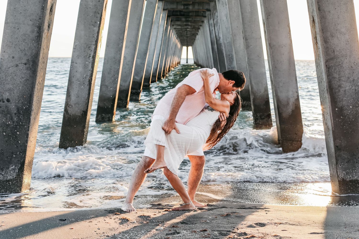 A couple in white outfits poses under a pier during sunset at the beach, creating romantic silhouettes.