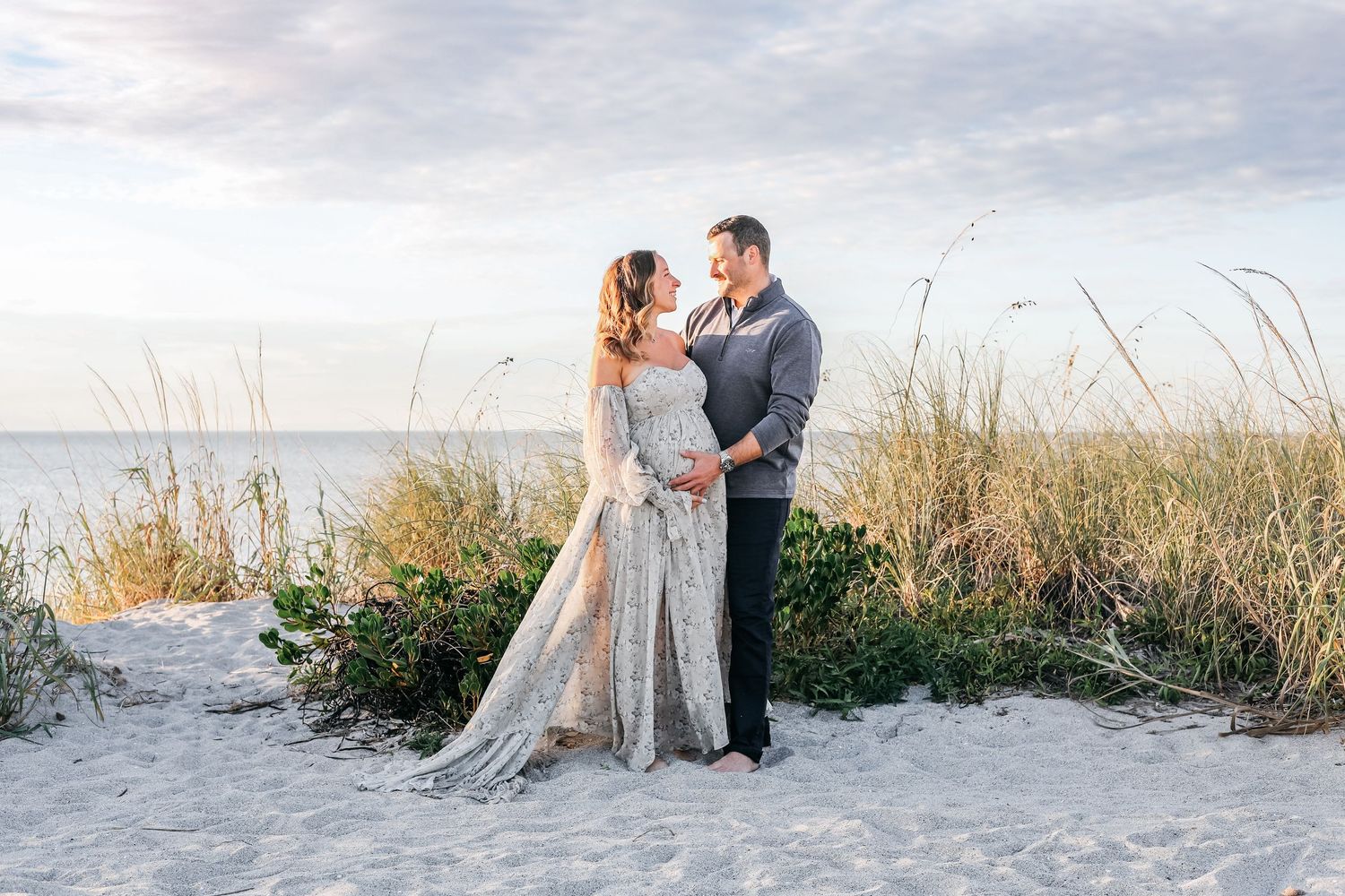 A couple in wedding attire shares romantic moments along a beach with sea grass and cloudy skies in the background.
