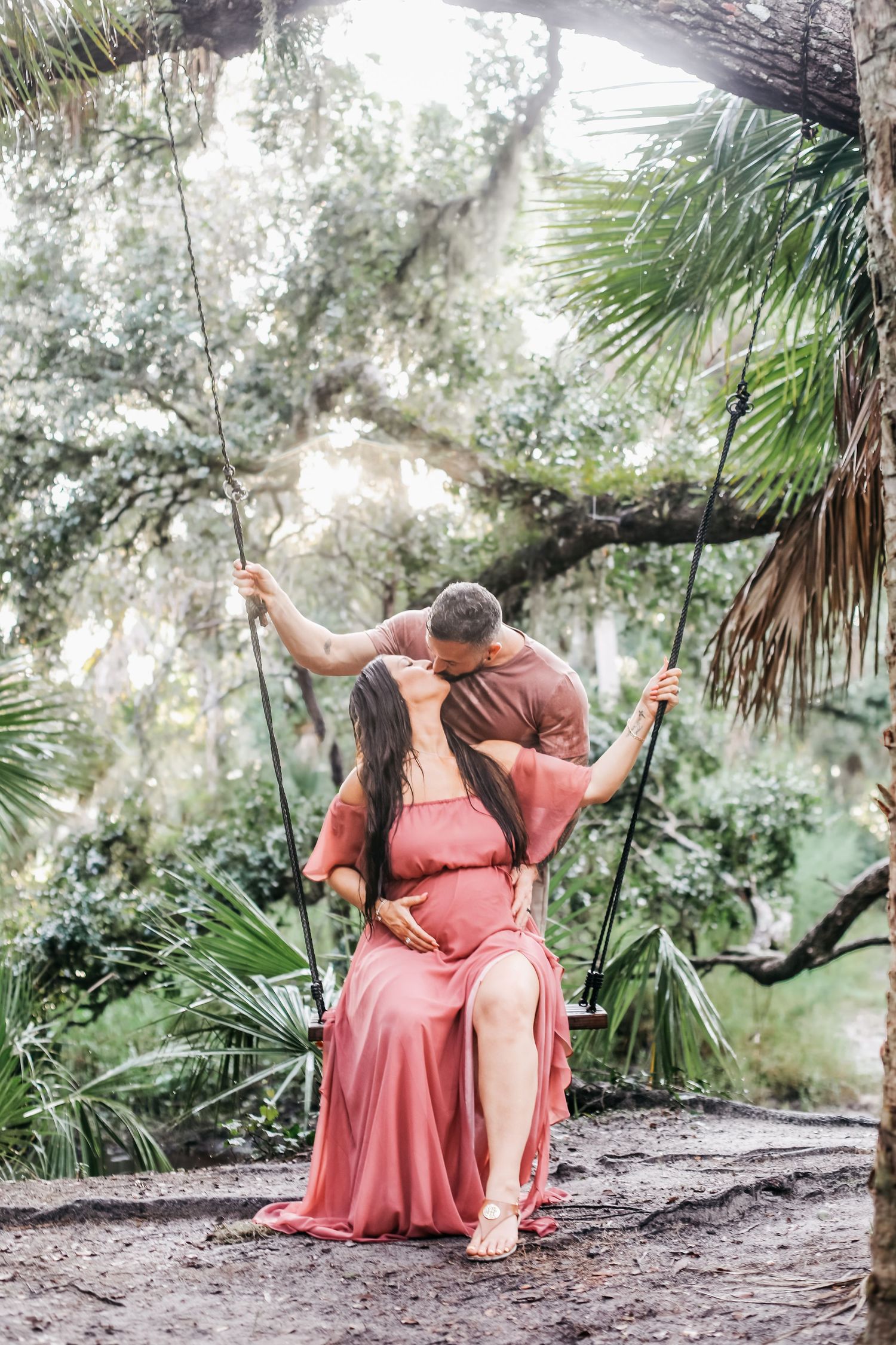 Two people in coral dresses sit together on a wooden bench in a lush tropical garden setting.