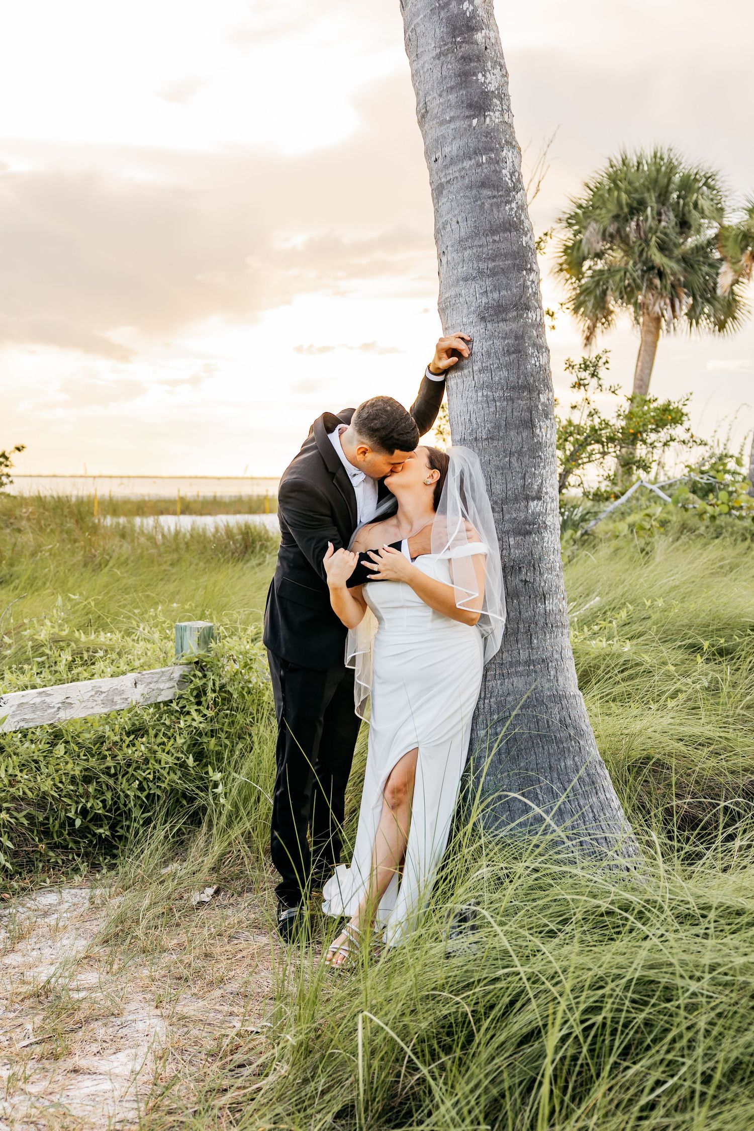 Bride and groom share a romantic kiss leaning against a palm tree at golden hour on a Florida beach wedding.