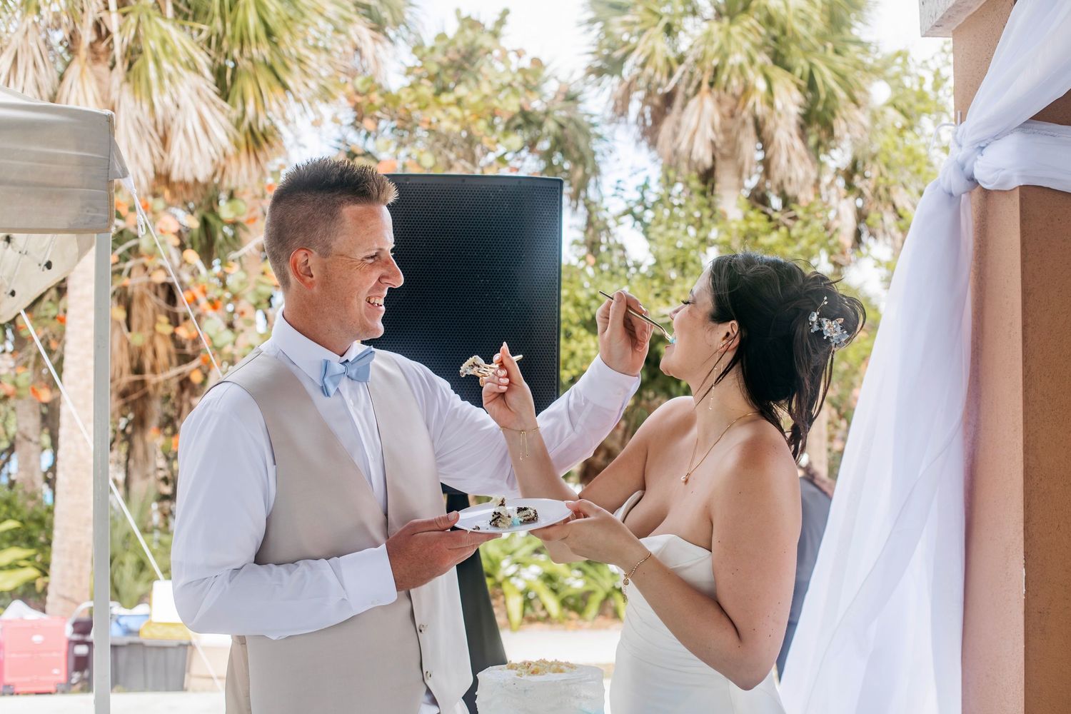 Bride and groom feeding each other wedding cake outdoors at a beach ceremony surrounded by palm trees.