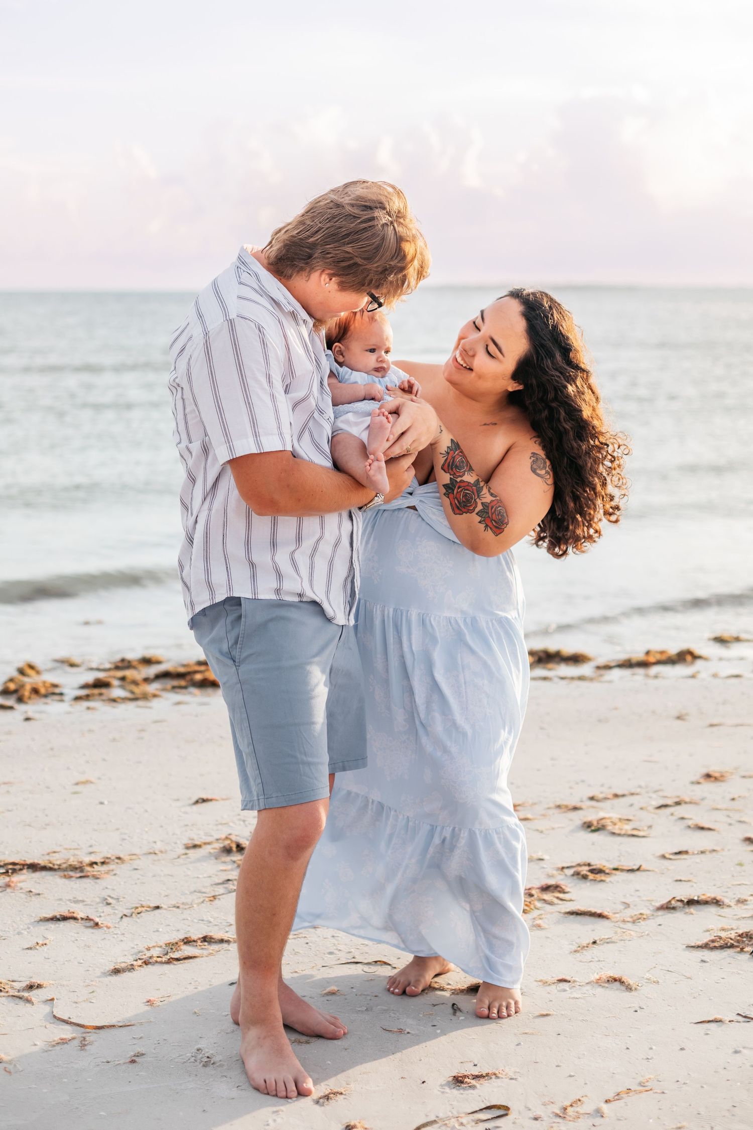 A couple stands barefoot on a sandy beach, lovingly gazing at their newborn baby near the ocean.