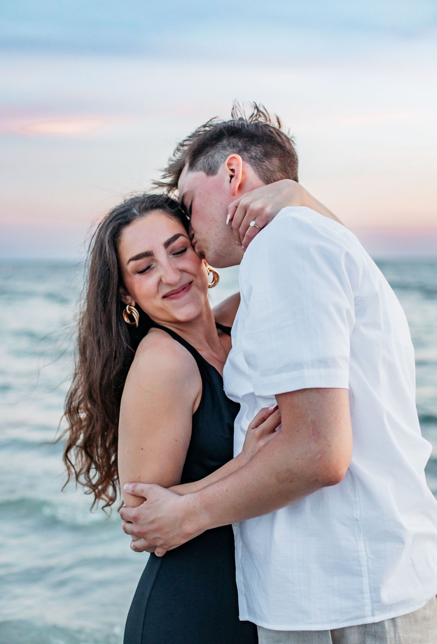 A couple embraces romantically on a beach at sunset, woman in black dress smiling as man kisses her cheek.