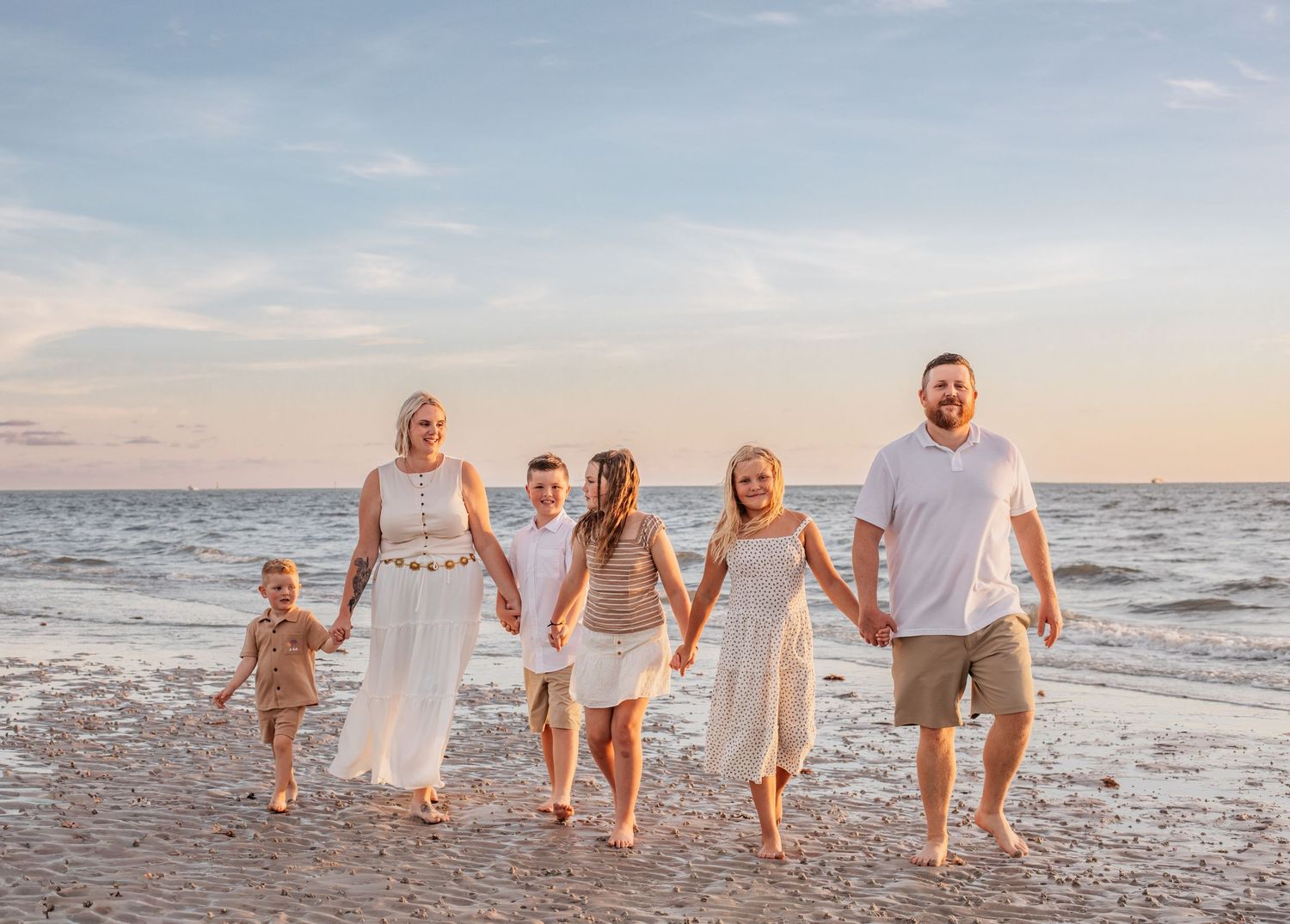 Family of six holding hands walking on a sandy beach at sunset in white and neutral outfits.
