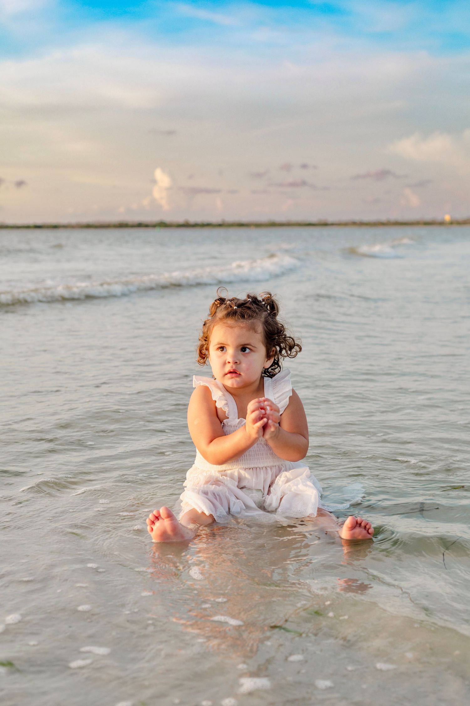 Small child sits in shallow ocean water wearing white at sunset.
