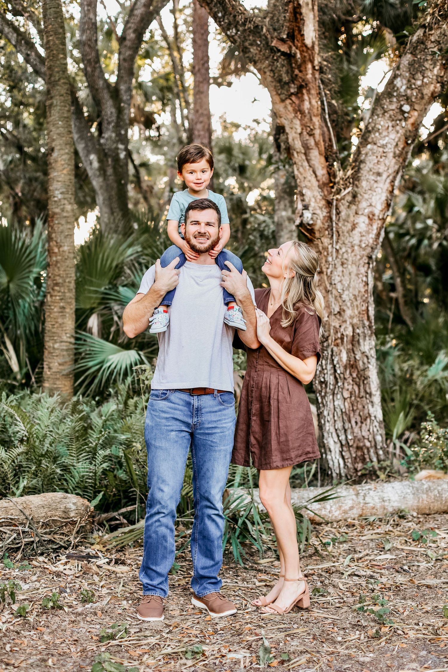 A couple stands together playfully in a wooded outdoor setting with palm trees and natural scenery.