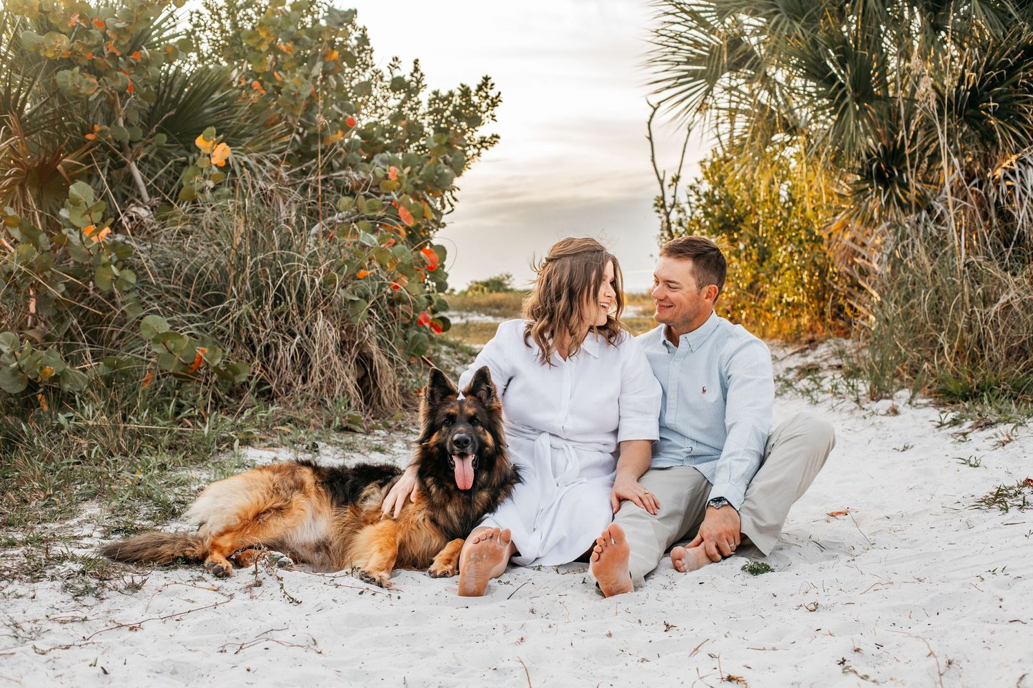 Couple sitting on white sand beach with German Shepherd dog, surrounded by lush greenery at golden hour.