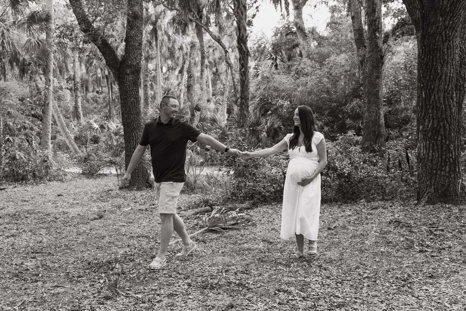 Black and white photo of a pregnant couple holding hands in a lush forest setting during a maternity photoshoot.