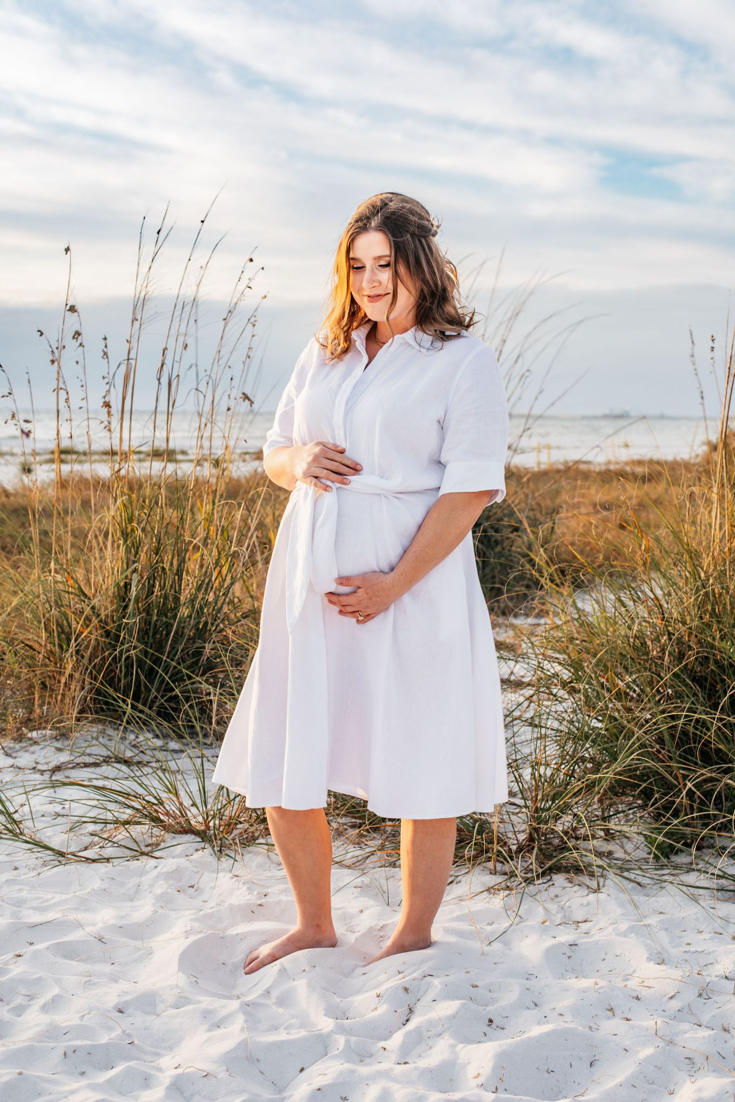 Pregnant woman in white dress standing barefoot on sandy beach with sea oats and ocean in background.