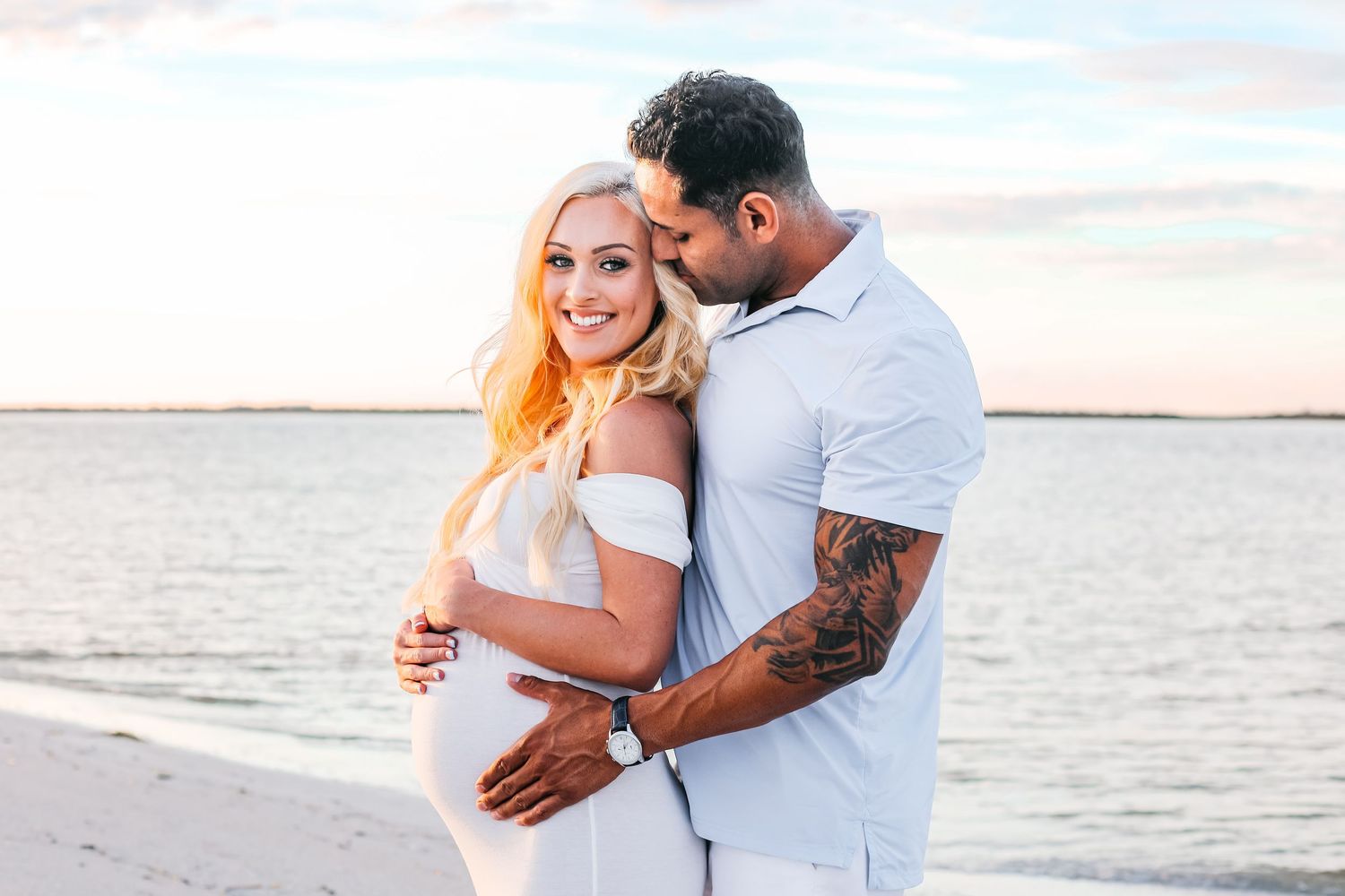 A couple embraces on a beach at sunset while wearing white and casual attire.