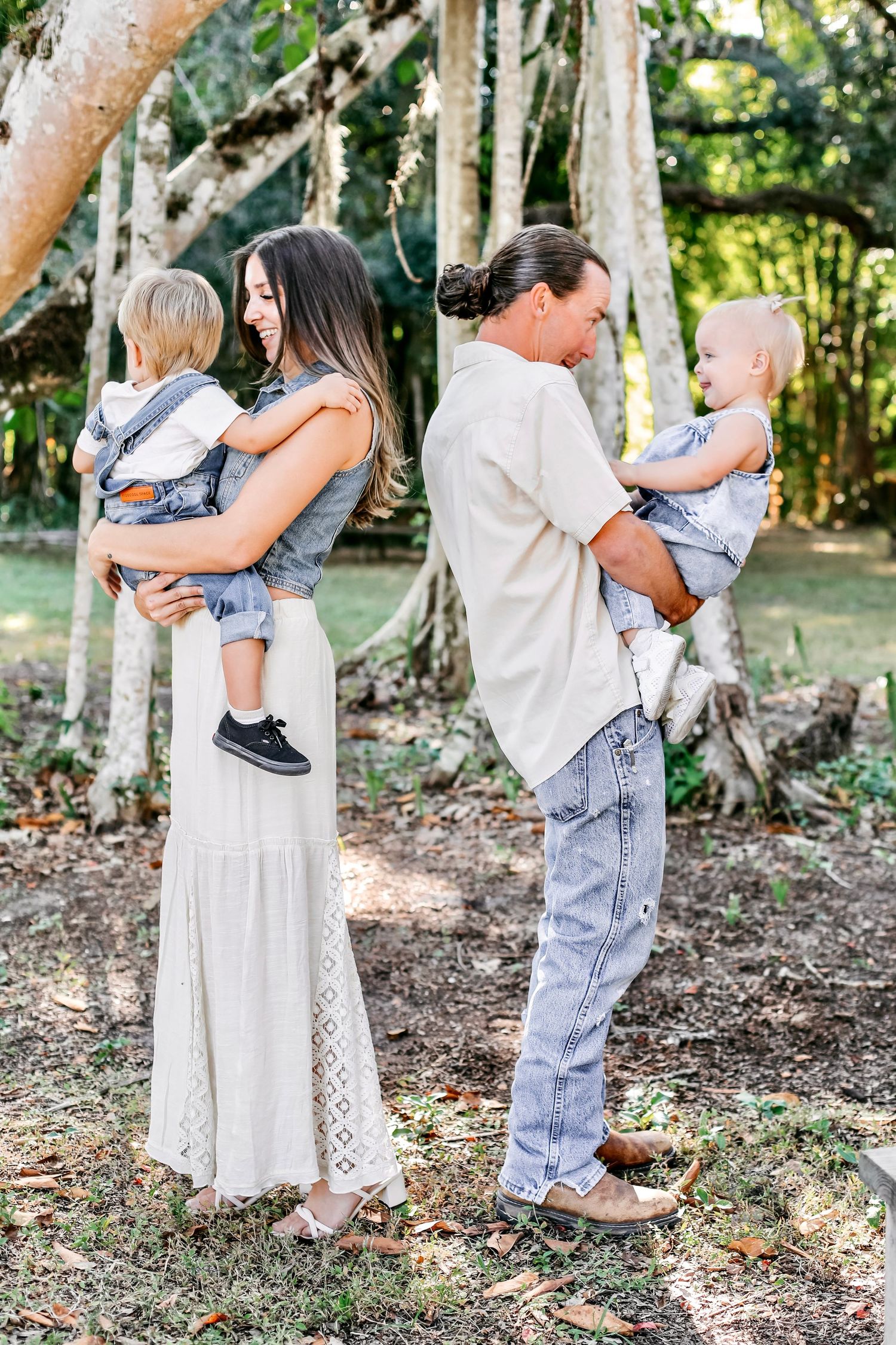 Family standing together outdoors in a wooded area wearing casual clothing and holding young children.