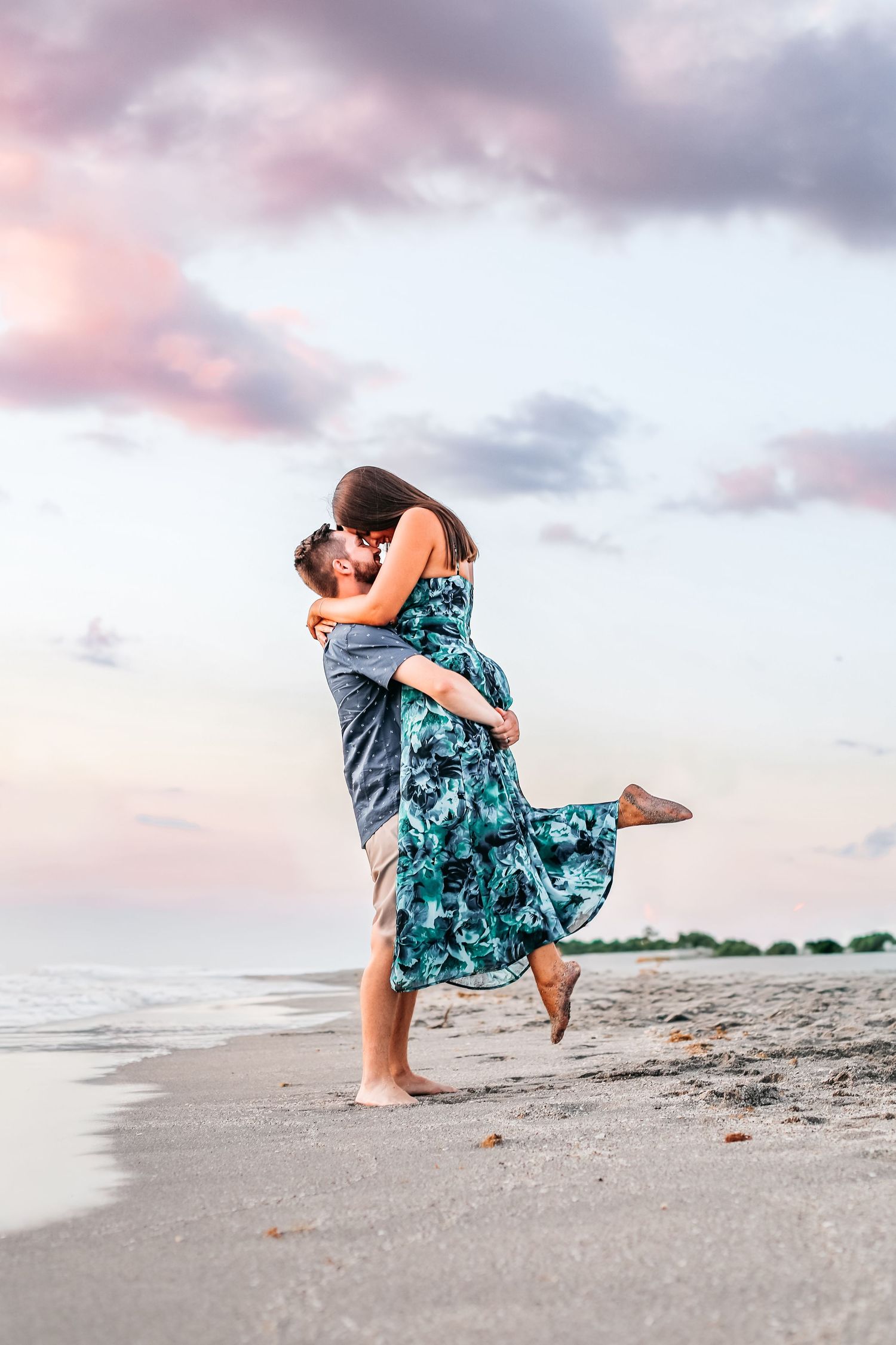 A couple embraces on a beach at sunset, with the woman wearing a flowing teal dress as they share a romantic moment.