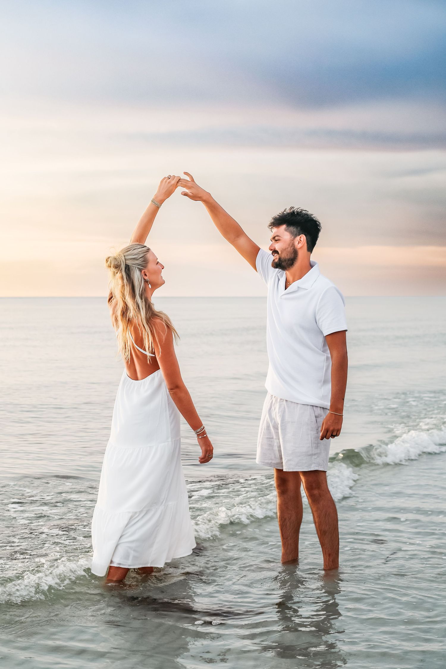 Couple wearing white outfits stands in shallow ocean water during sunset creating artistic poses together.