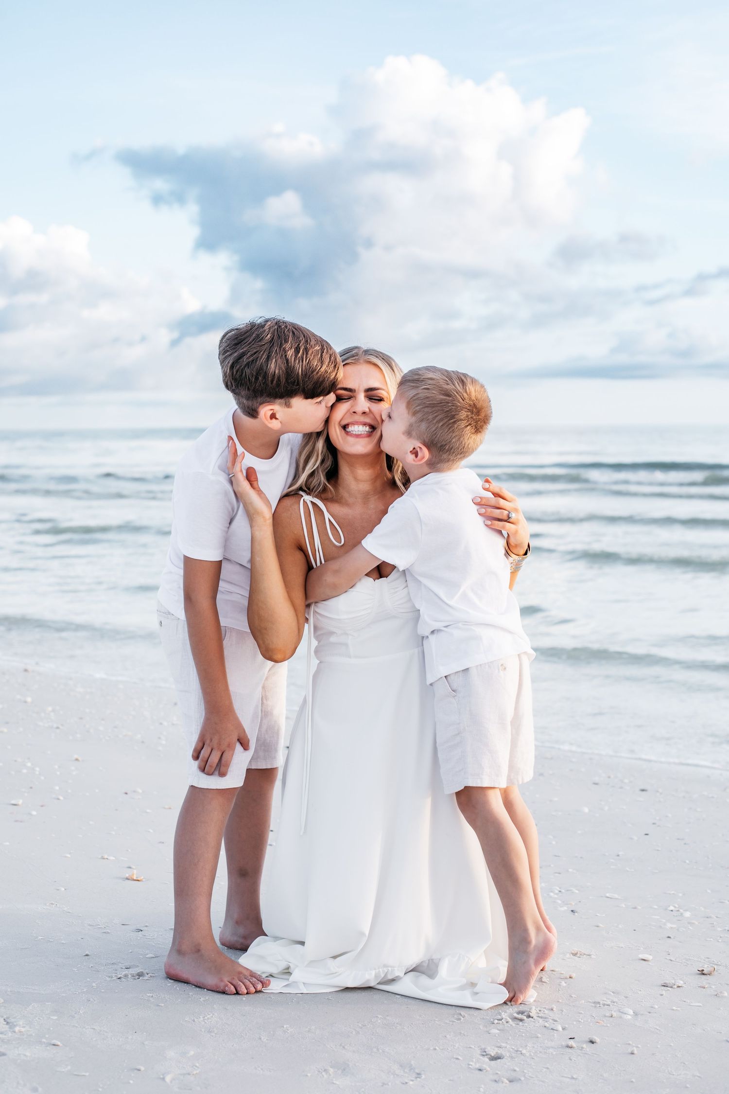 A family wearing white outfits shares tender moments together on a sunny beach with ocean waves in the background.