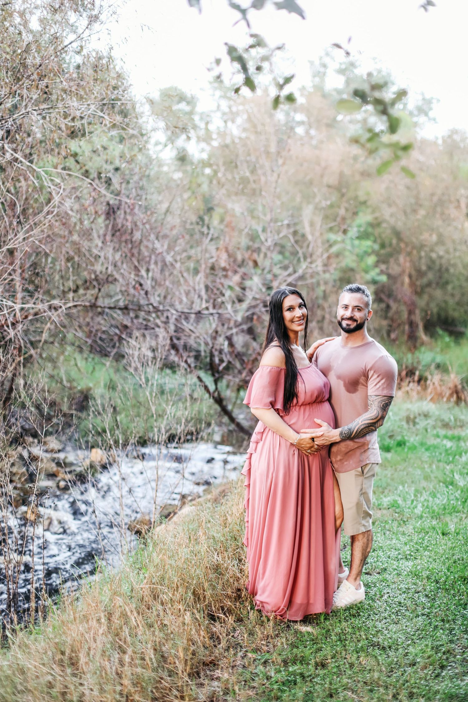 A couple poses together outdoors in coordinated outfits by a stream with pink dress and tan shirt against natural scenery.