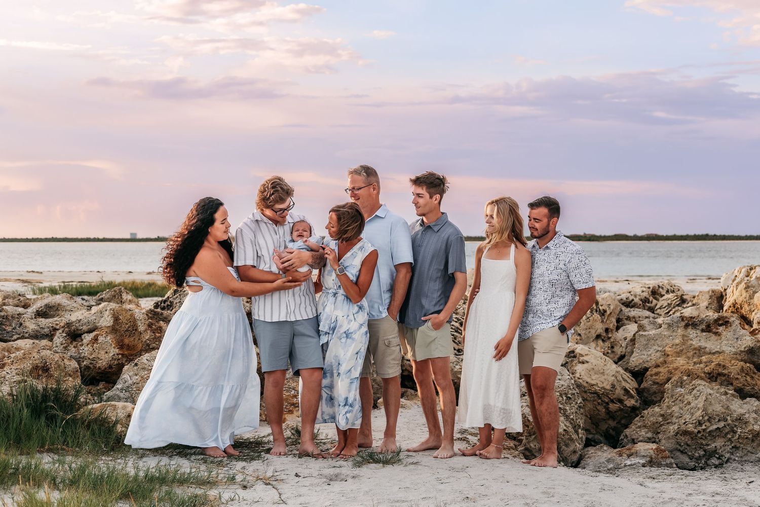 Family group portrait on rocky beach at sunset, pastel sky backdrop, adults and children in coordinated outfits.