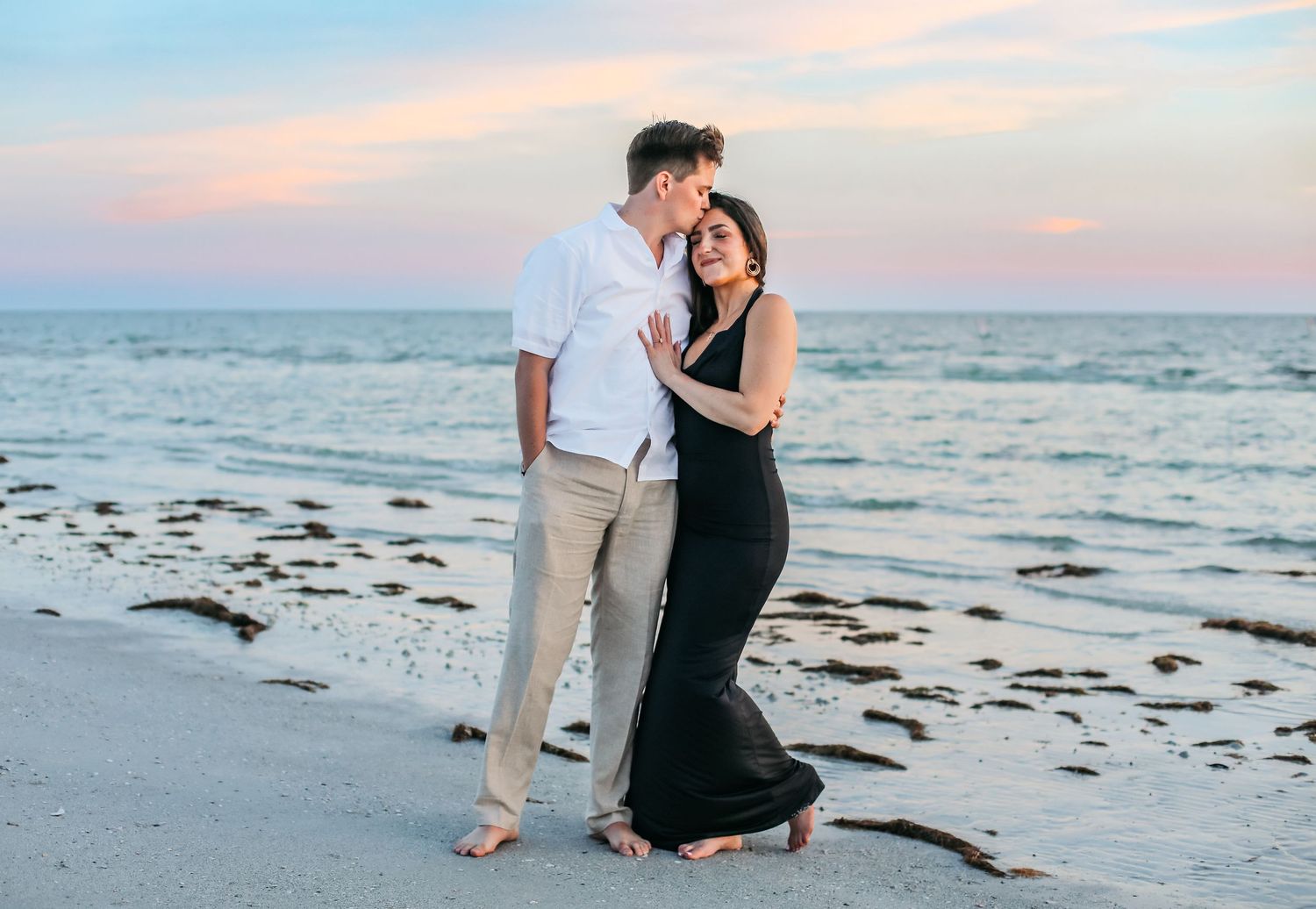 Couple embracing on a sandy beach at sunset, woman in black gown, man in white shirt, ocean backdrop.