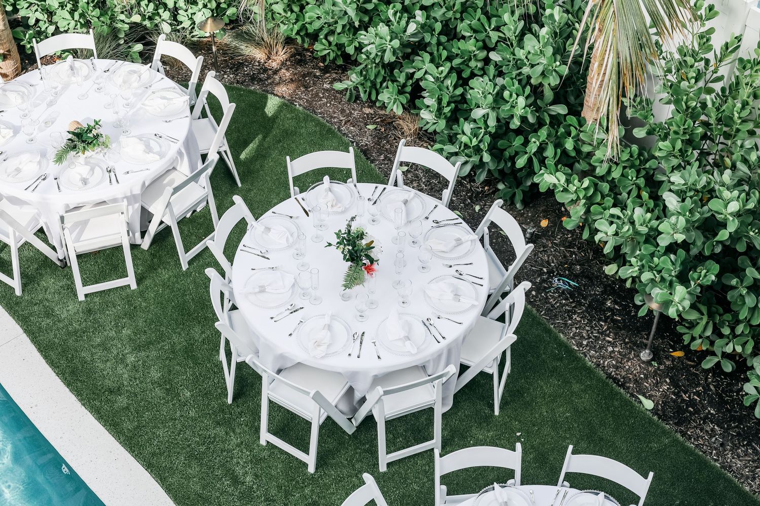Elegant outdoor wedding reception with round white tables and folding chairs on green lawn beside pool.
