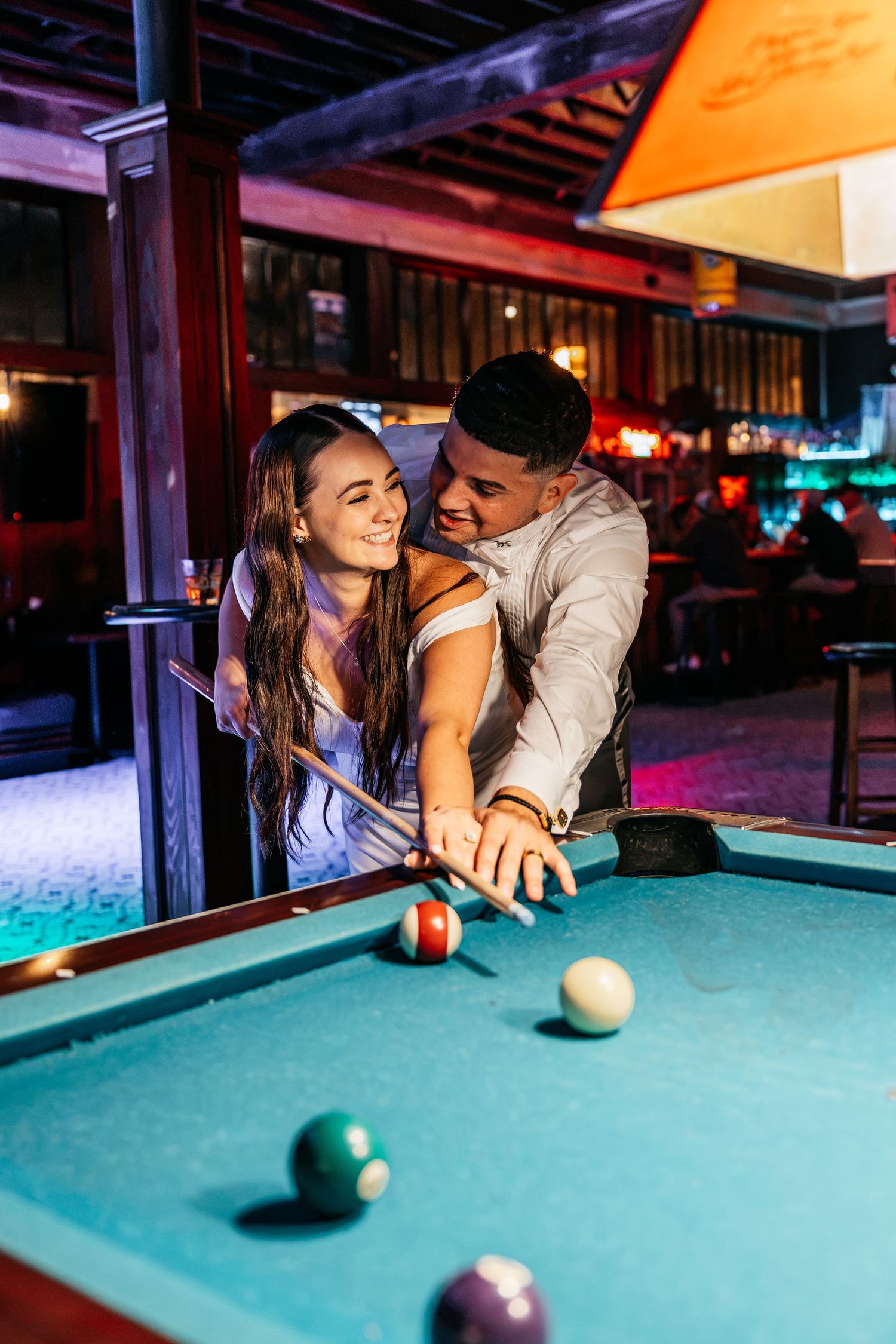 A couple smiles while playing pool together at a vibrant bar, the man guiding the woman as she holds a cue stick.