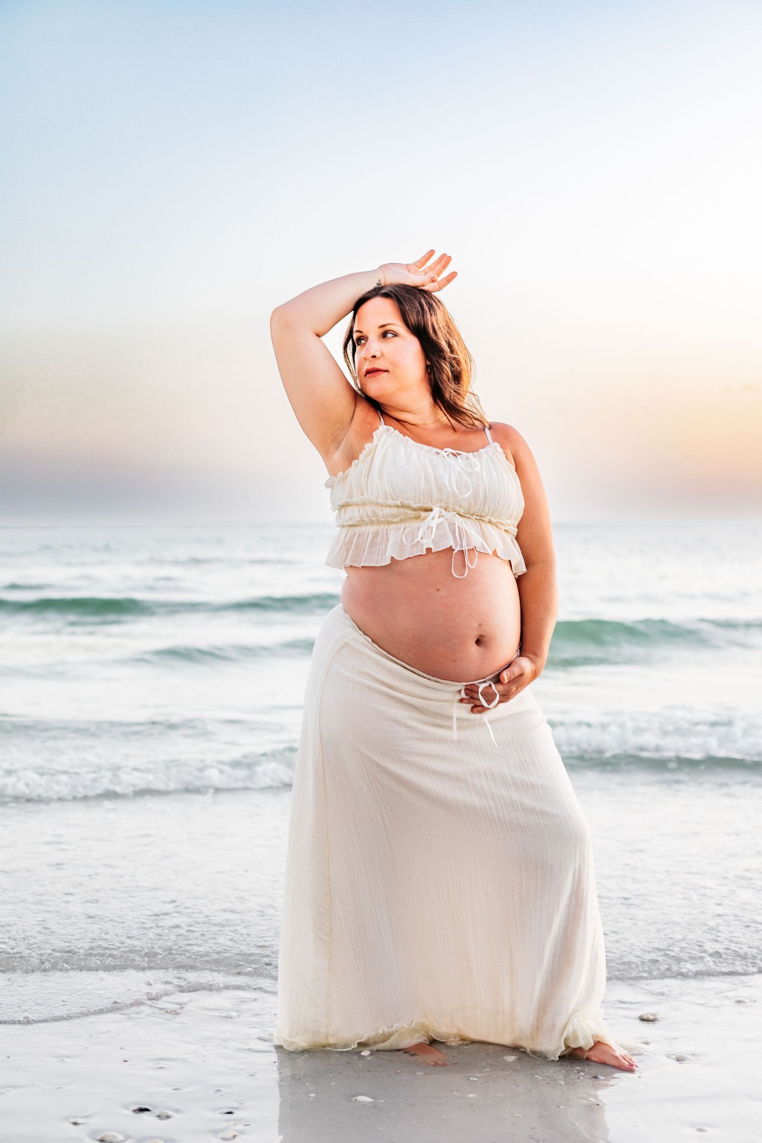 Expecting mother in flowing white dress poses gracefully on beach at sunset with ocean waves in background.