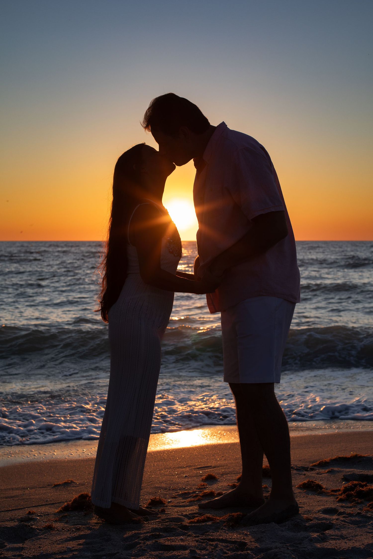 Silhouetted couple shares a romantic kiss on the beach at sunset with ocean waves in the background.