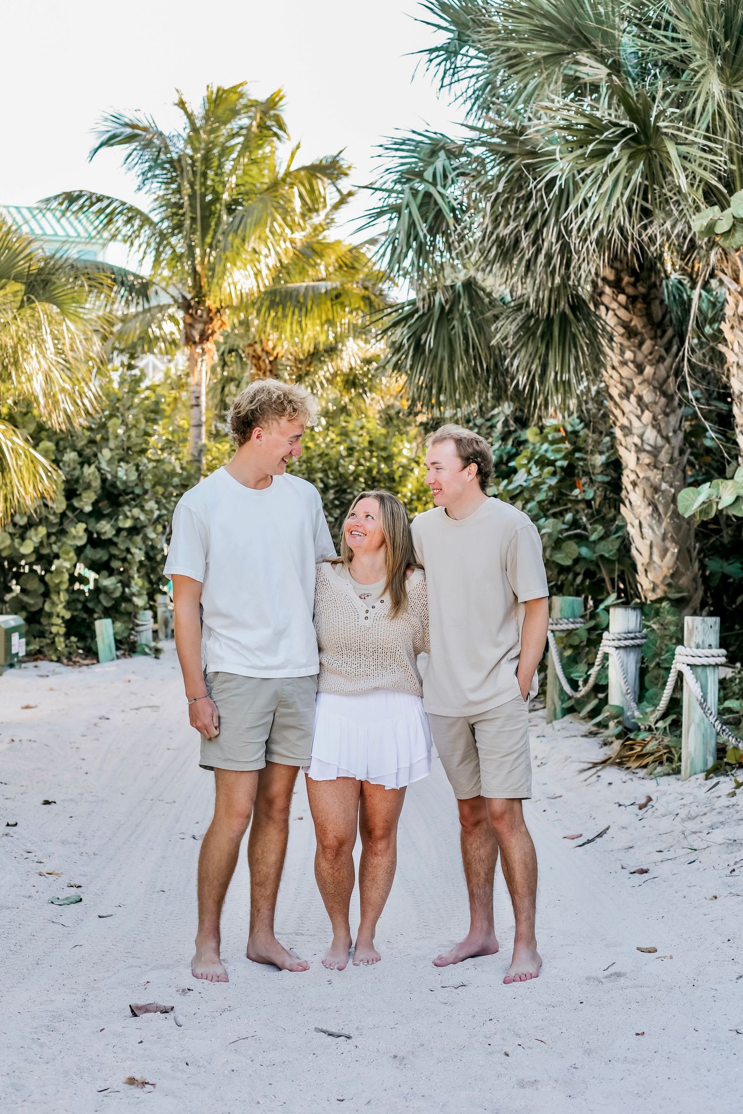 Family in coordinating white and beige outfits poses together on a tropical beach with palm trees in the background.