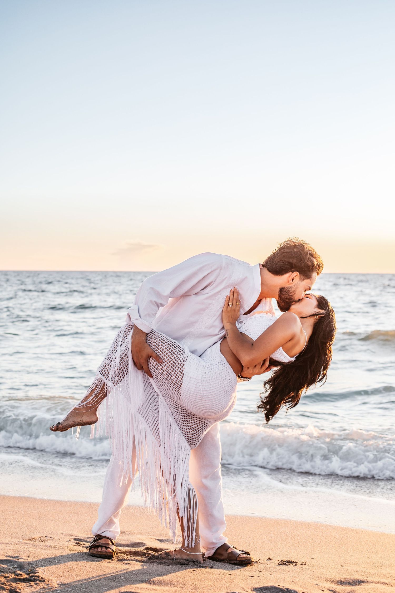 Romantic couple in white outfits dancing on a beach at sunset with ocean waves in the background.